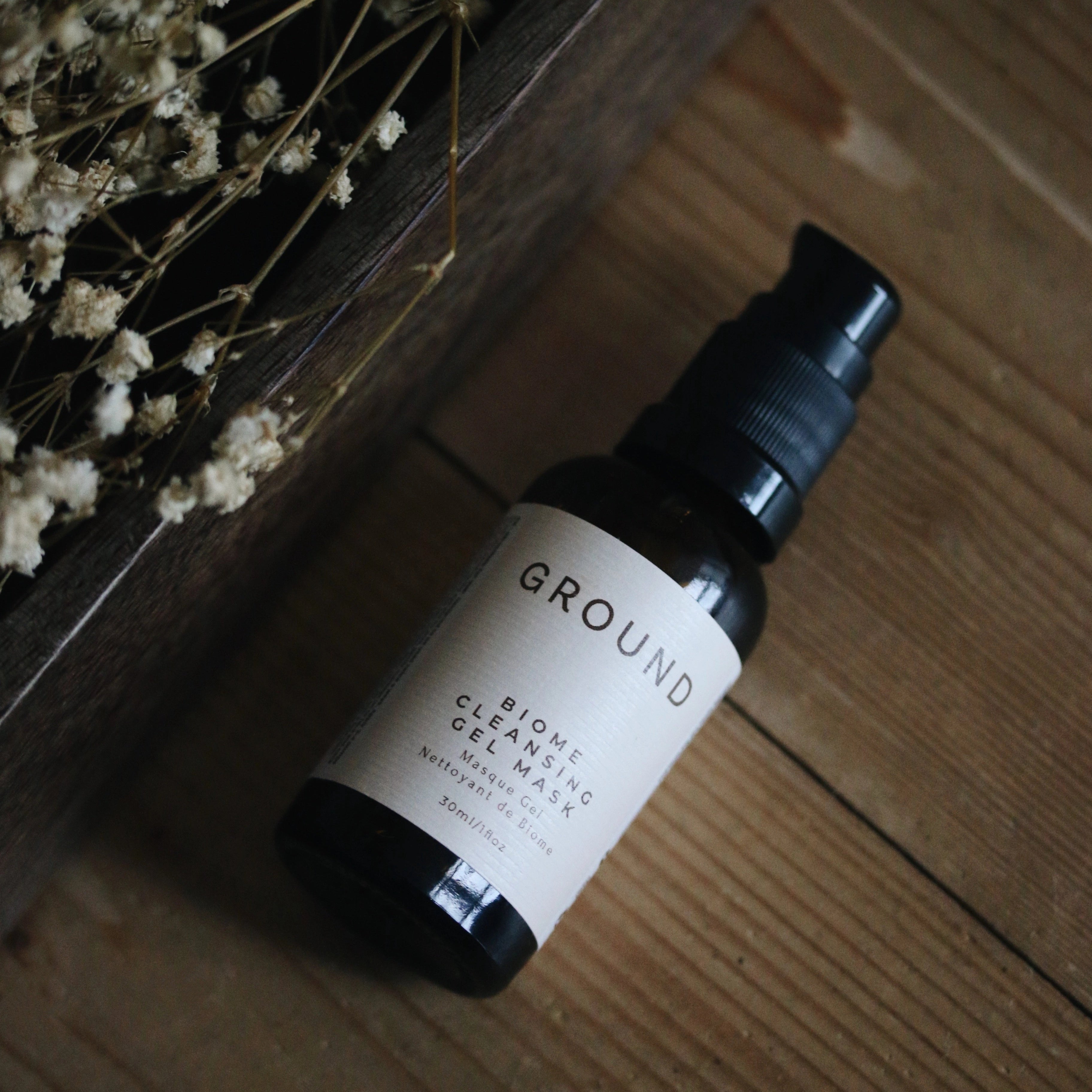 Photograph of bottle of Ground Biome Cleansing Gel Mask in 30ml size lying on a wooden countertop with white flowers called baby's whisper next to it.