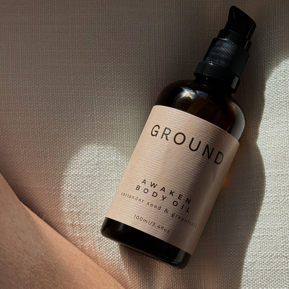 A 100ml amber glass bottle of GROUND Awaken Body Oil with a black pump and a beige label, resting on a textured beige fabric surface with a warm shadow.