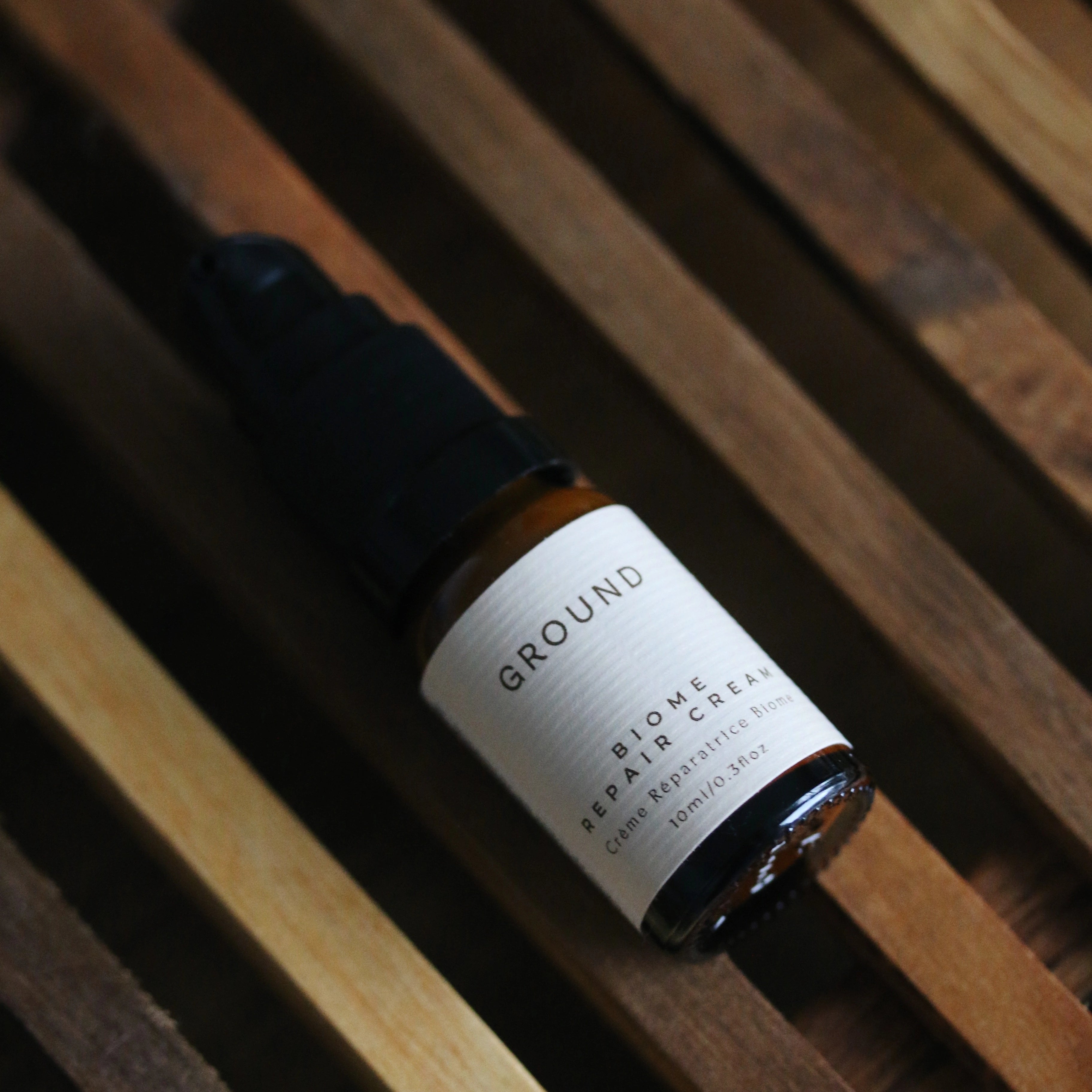 Image of a brown glass bottle with pump dispenser lying flat on a wooden rack on top of a wooden surface. It is a 10ml bottle.