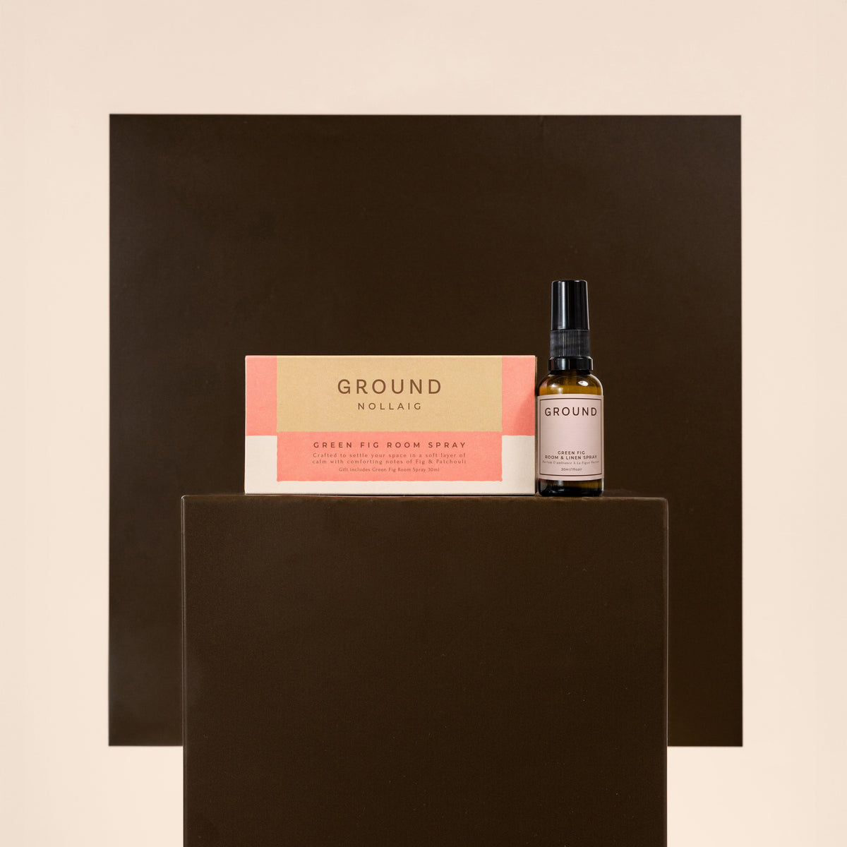 A small amber spray bottle and its matching box labeled GROUND Green Fig Room Spray, displayed on a dark brown pedestal against a warm beige background.