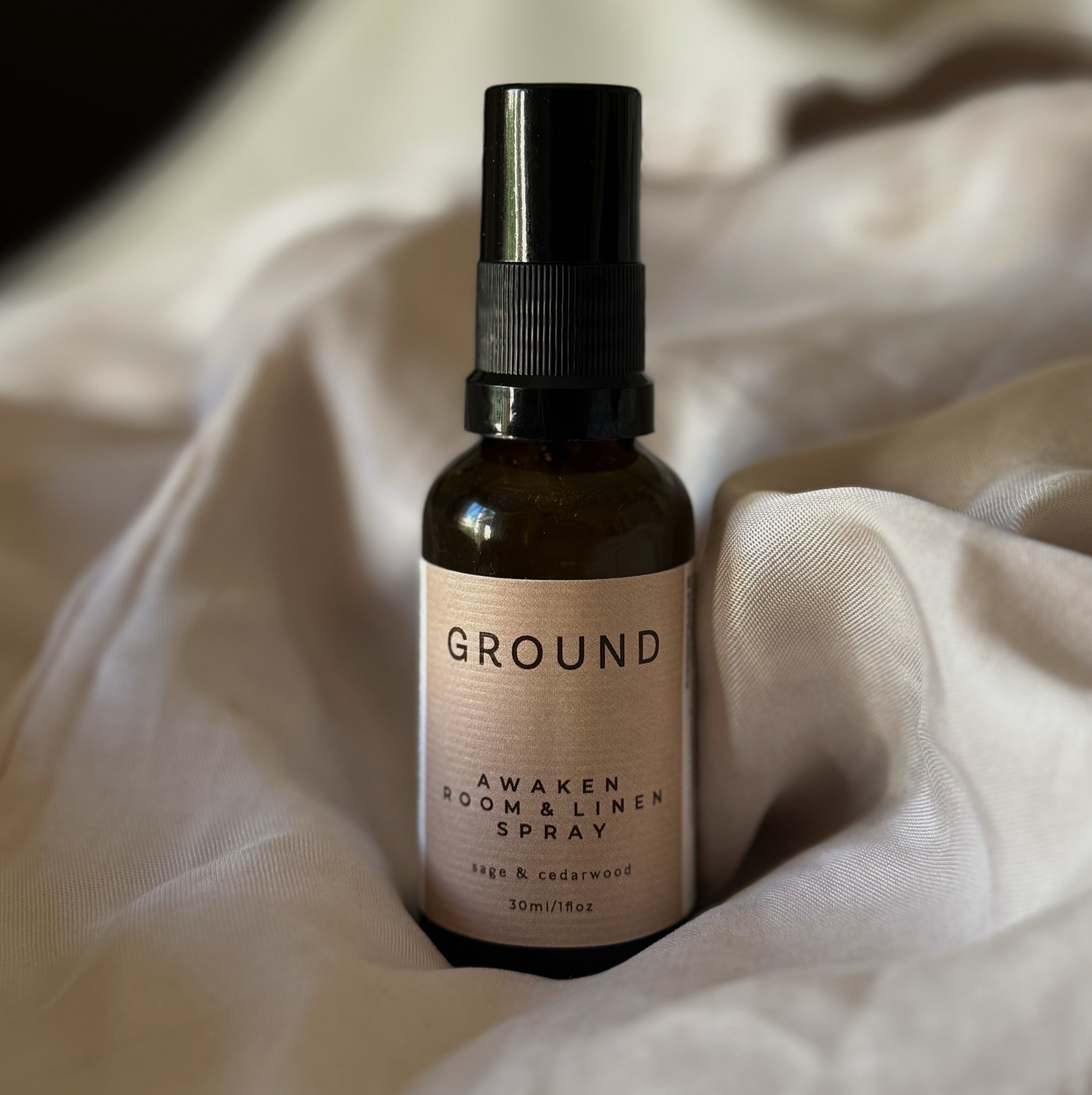 Image of a brown glass bottle with a black lid labelled with a beige labelling stating Awaken Room & Linen Spray Sage & Cedarwood - 30ml. Surrounded by neutral material creating a soft backdrop.