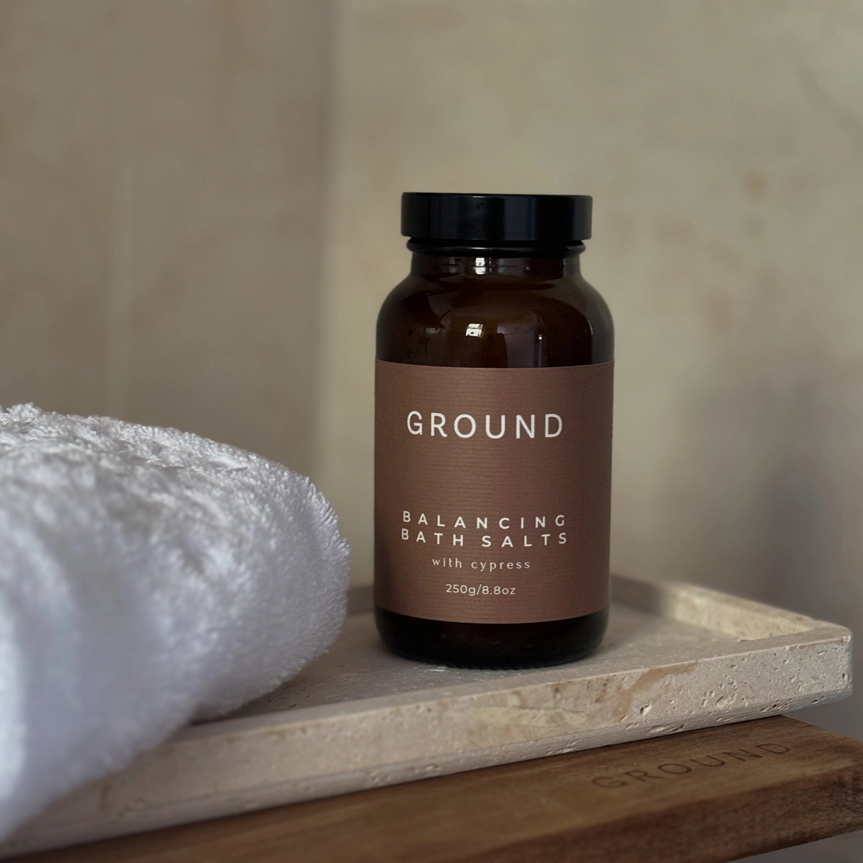 Photo of a jar labelled Ground Balancing Bath Salts with cypress 250g. Displayed on a stoneware tray beside a white towel on top of a GROUND branded wooden board.
