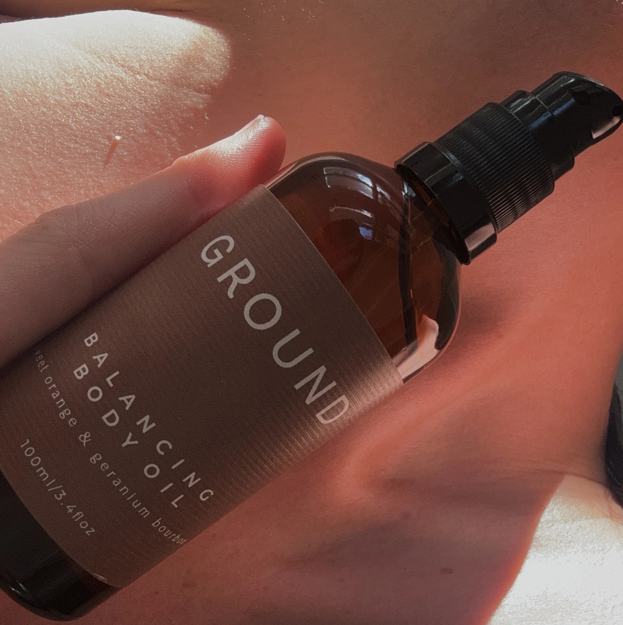 Image of a glass bottle of body oil being held up against the neckline. It is a brown glass pump bottle containing 100ml of a sweet orange and geranium bourbon fragranced oil.