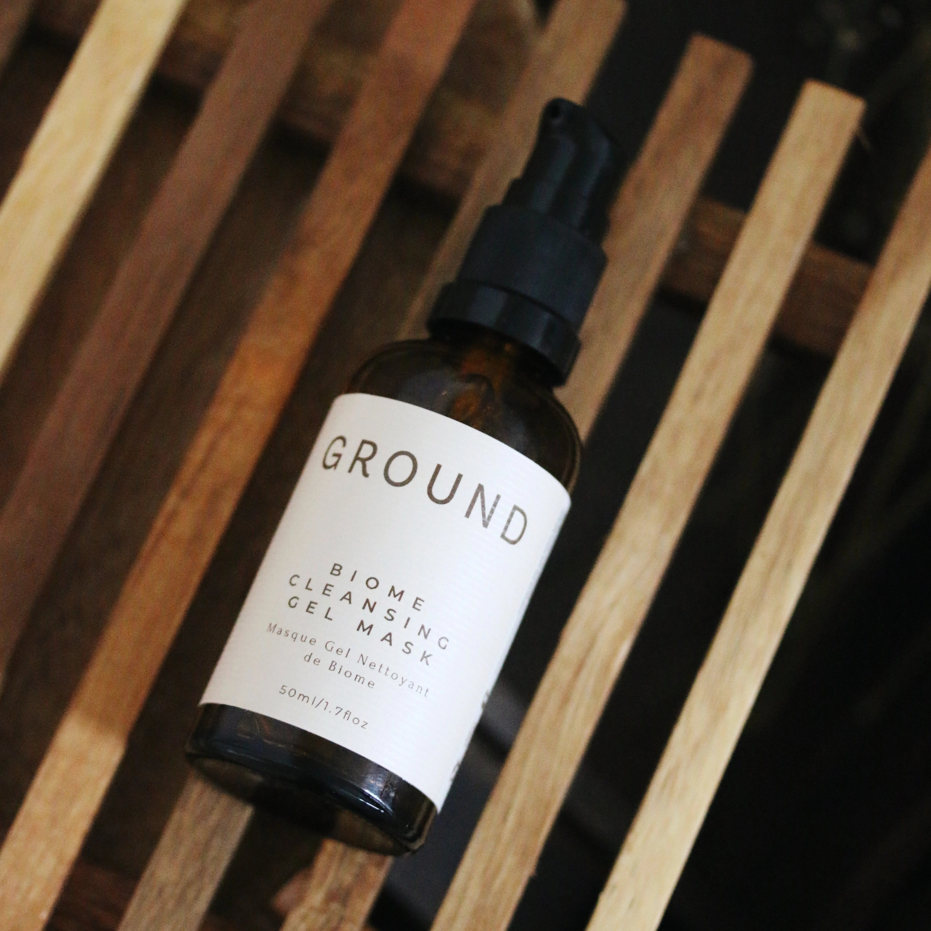 A bottle of BIOME Cleansing Gel Mask by GROUND lying on a wooden rack. The label and packaging are both beige in colour with brown writing showing a 100ML bottle.