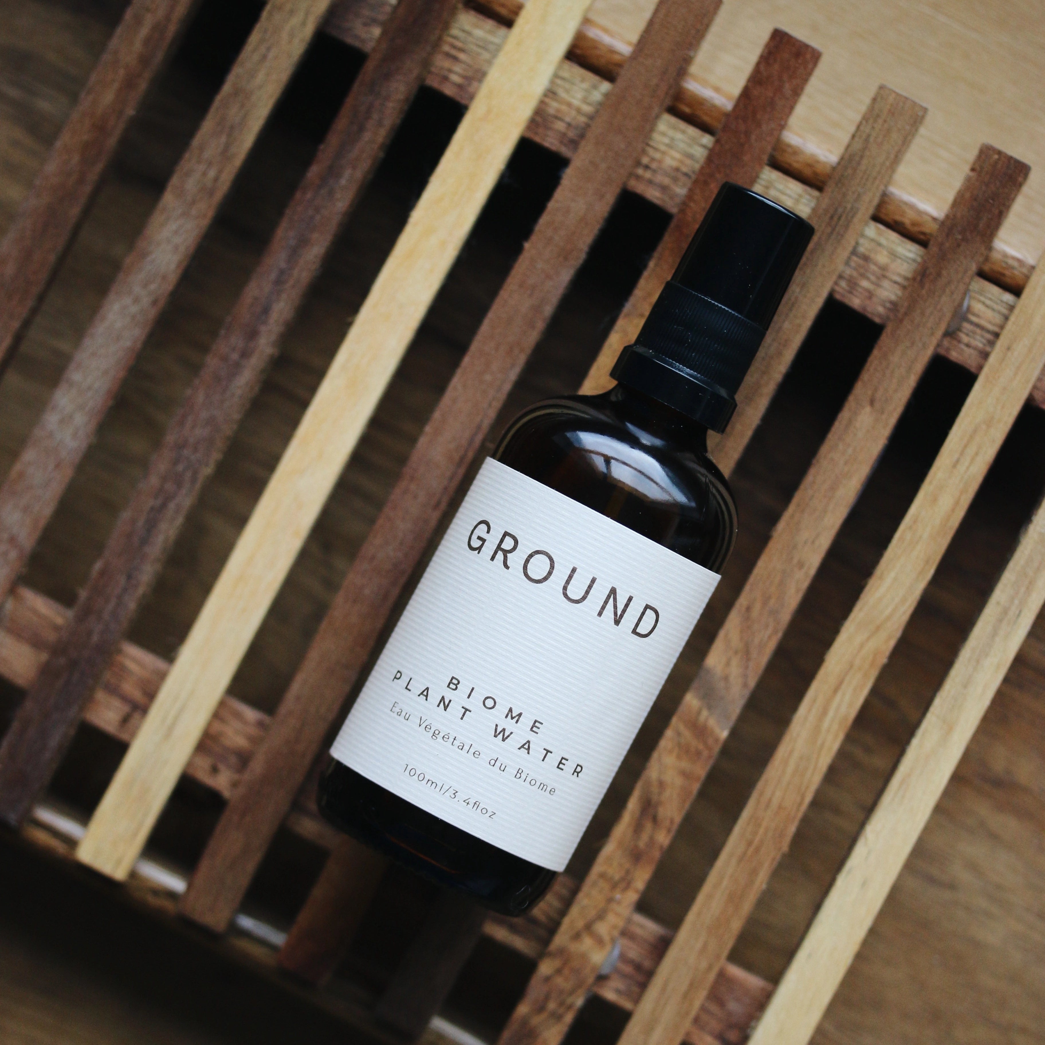 Image of a bottle of Ground Biome Plant Water. It is a glass bottle with a spray dispenser top, lying on its back on a wooden rack over a wood surface.
