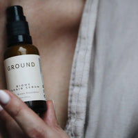 Photo of a bottle of Ground Biome Repair Serum being held up in front of a blurred background.
