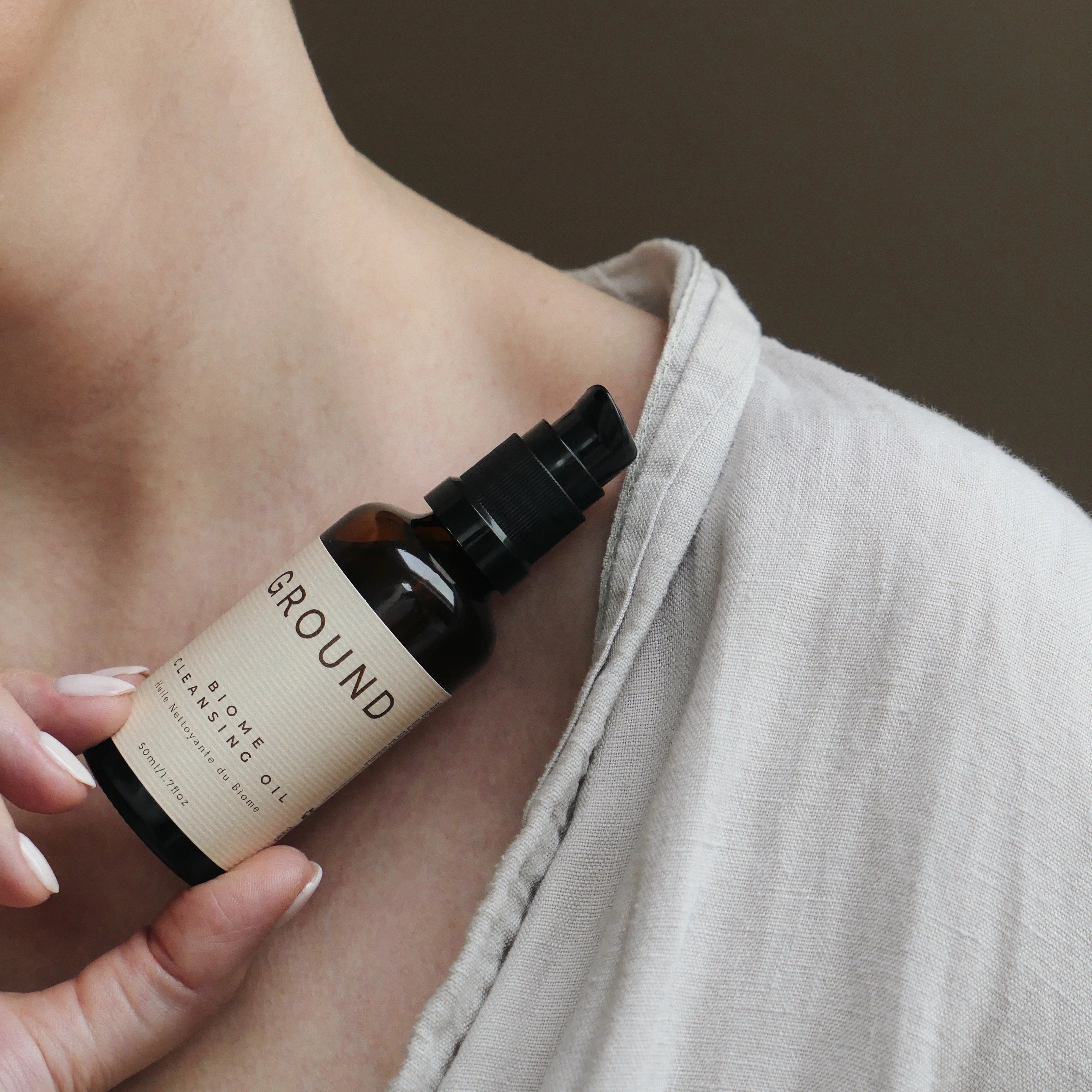 Photograph of a bottle id Biome cleansing oil being held up to a person's neck. They are wearing neutral colours against a dark green background.