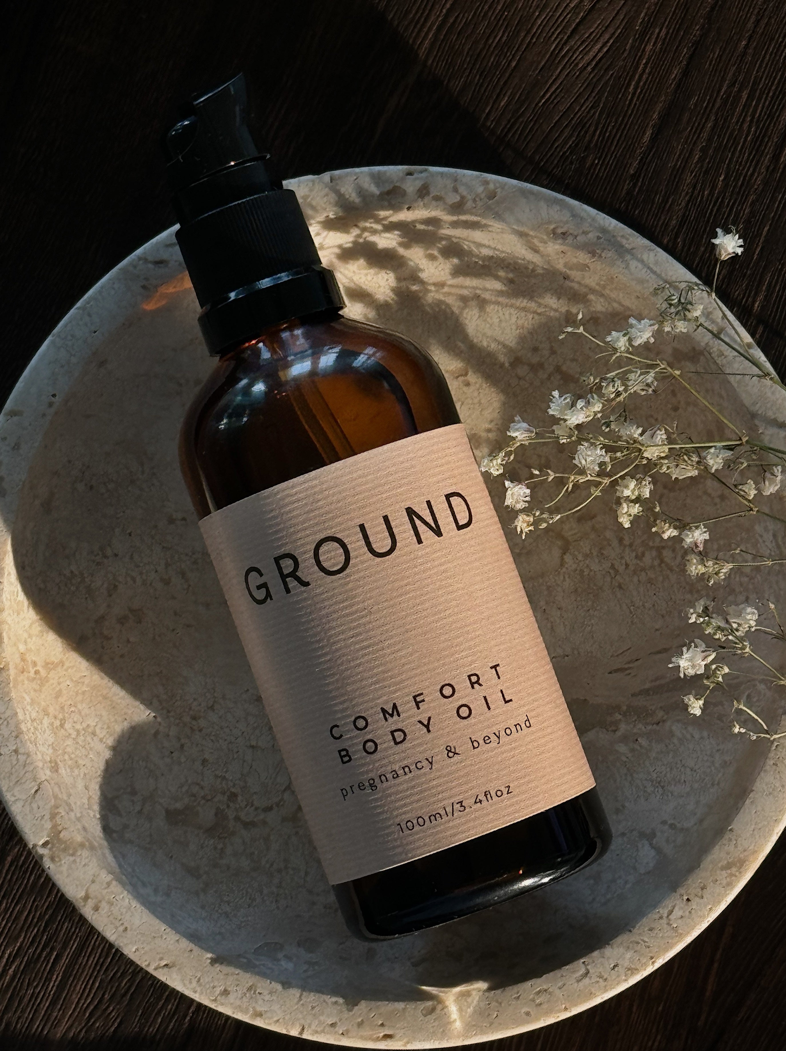 Photograph of a pump bottle of Ground Comfort Body Oil for pregnancy and beyond in 100ml size. It is balanced on its back in a stone dish with small dried white baby's whisper flower on the side.