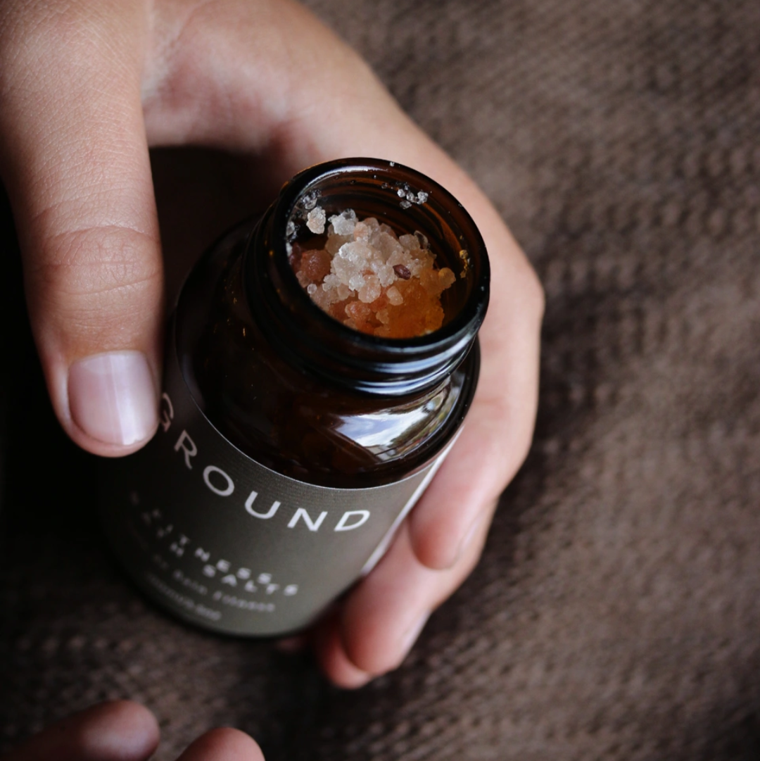 Hand holding a small brown bottle labeled 'Ground' on a deep brown label in front of a textured cloth surface.