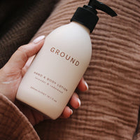 Hand with pale pink polished nails holding a 'GROUND' hand and body lotion, lavender and cedarwood bottle against a brown textured background.