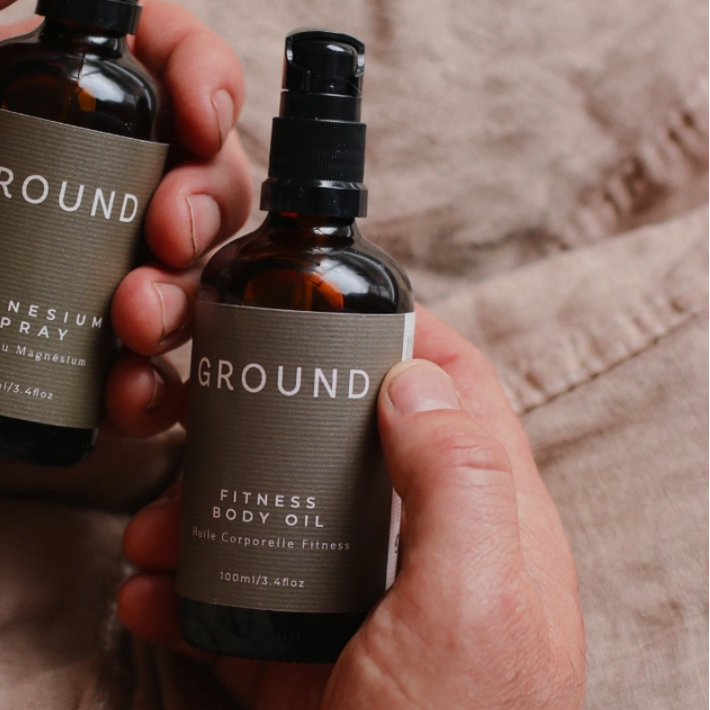 Two bottles of 'GROUND' products held by a person against a neutral linen background. One is Ground Magnesium Spray 100ml and the other a Fitness Body Oil 100ml / 3.4 fl oz.