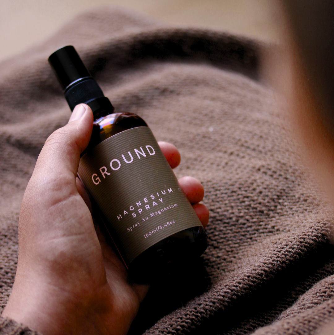 Hand holding a bottle of 'GROUND' magnesium spray in front of a textured brown surface.