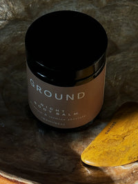 Photo of a pot of Ground Night Body Balm beside a Ground Gua Sha facial tool. The scene set is dimly lit against dark stone background.