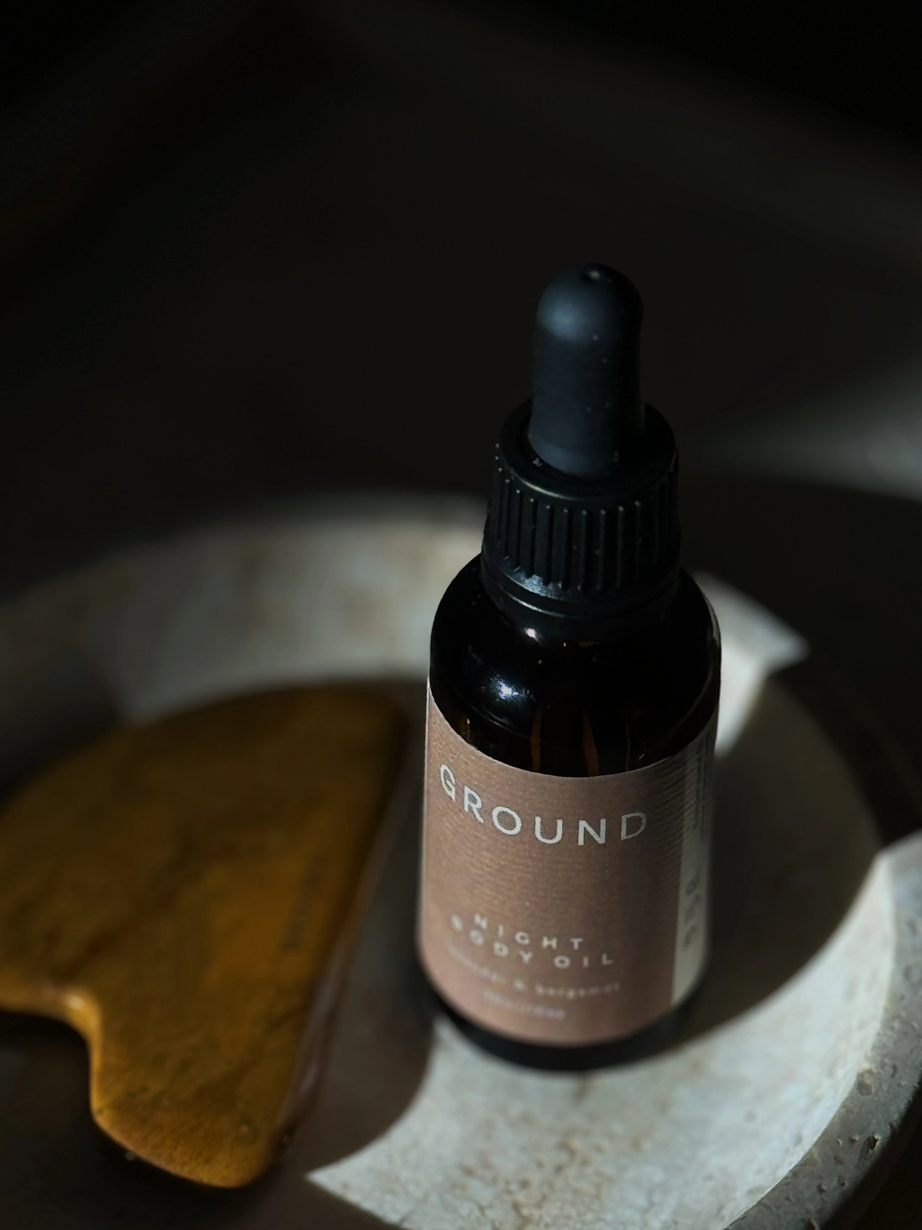 Photograph of a brown glass bottle with dropper lid sitting on a light stone dish beside a gua sha facial tool. The bottle is labelled as Night Body Oil.