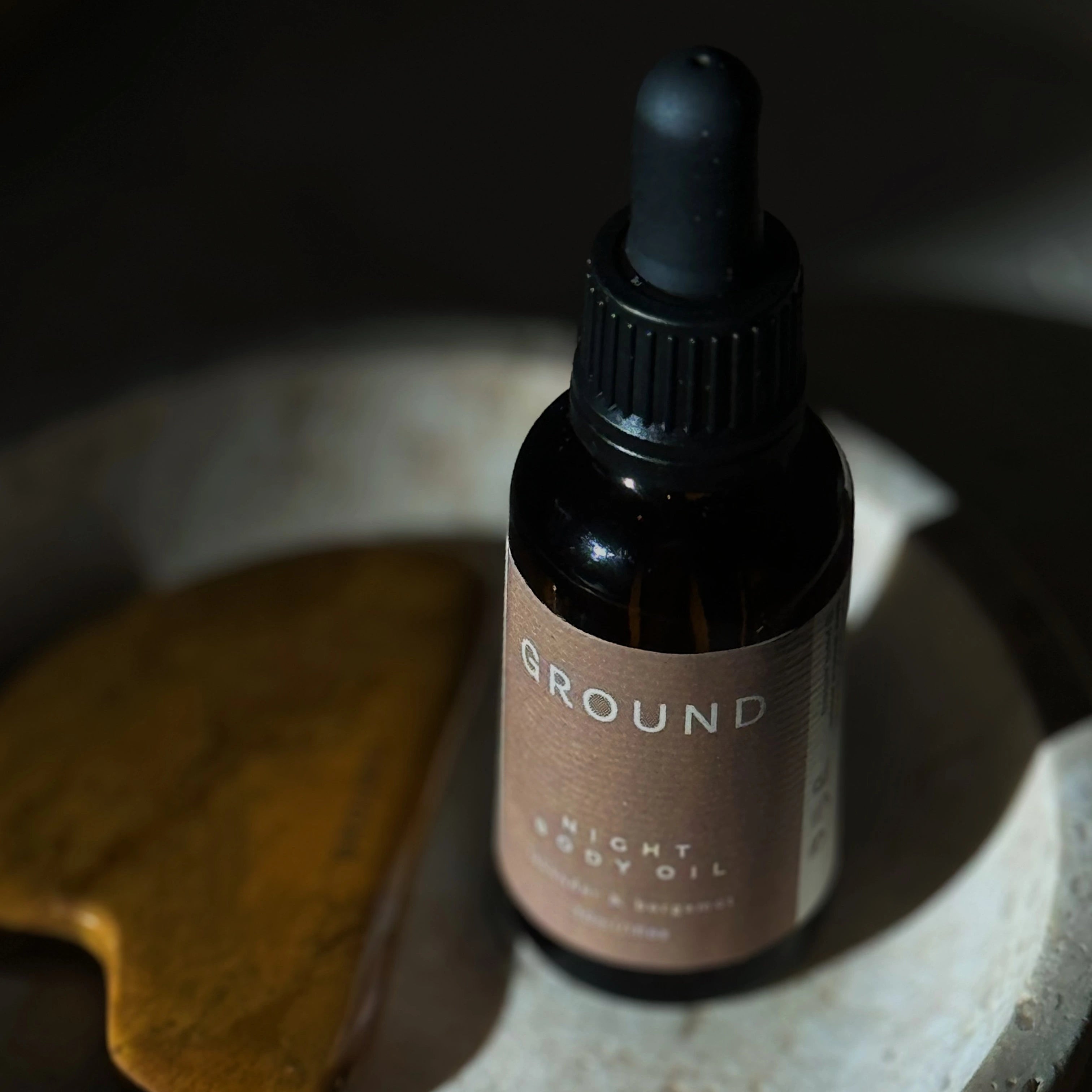 Photograph of a brown glass bottle with dropper lid sitting on a light stone dish beside a gua sha facial tool. The bottle is labelled as Night Body Oil.