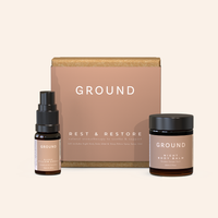 Ground skincare products sitting beside a cardboard gift box wrapped in branded light brown sleeve stating 'GROUND' Rest and Restore, natural aromatherapy to soothe and support. In the foreground there is a brown glass bottle of Ground Sleep Pillow Spray and a brown glass jar of Ground Night Body Balm. All sitting in on a white background.