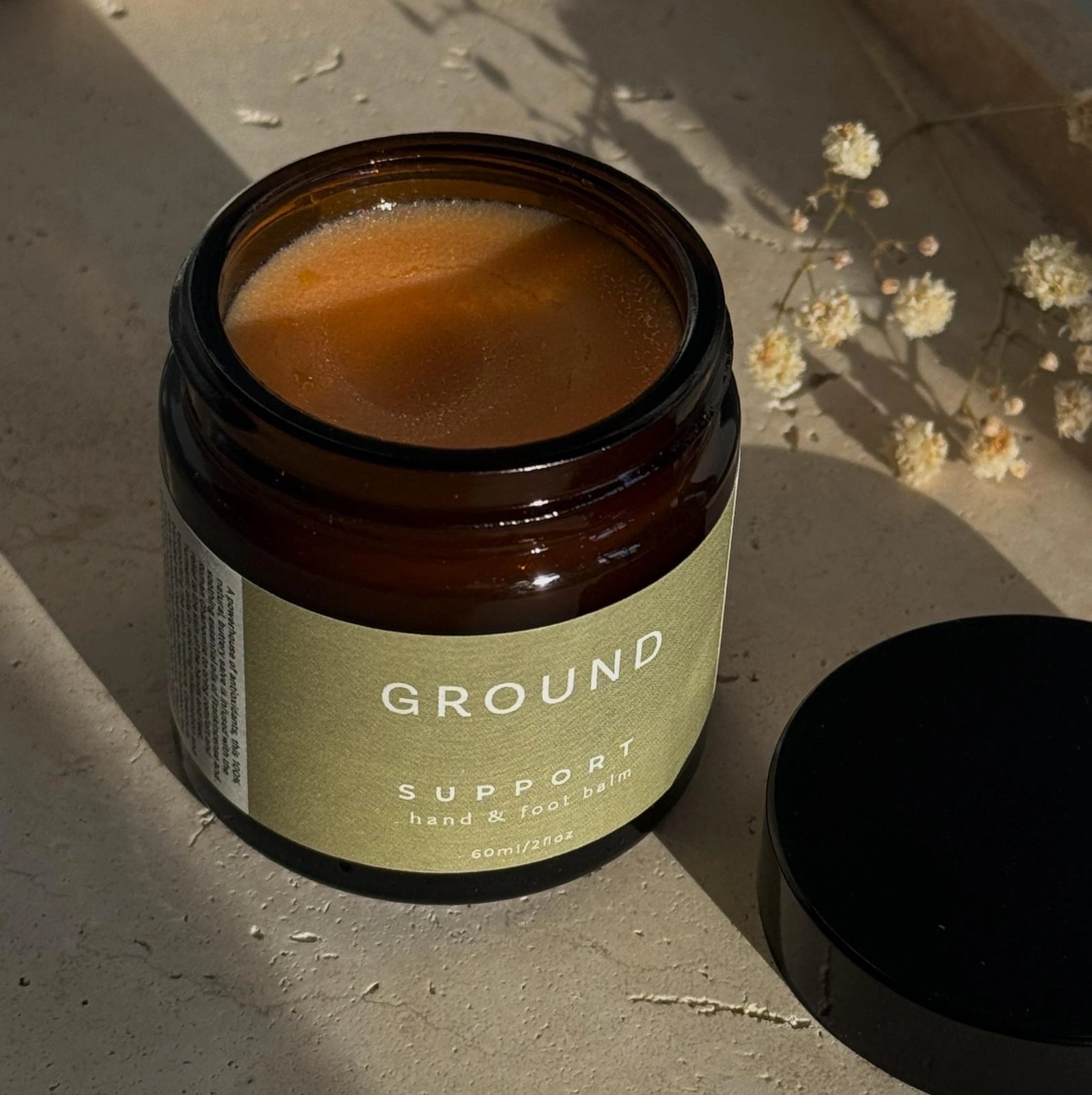 Photograph of a glass pot of balm with a sage green label with 'Support' hand & foot balm - 60ml and 2 fl oz. The photograph shows a jar with a sage green label sitting in the sun on a concrete surface.