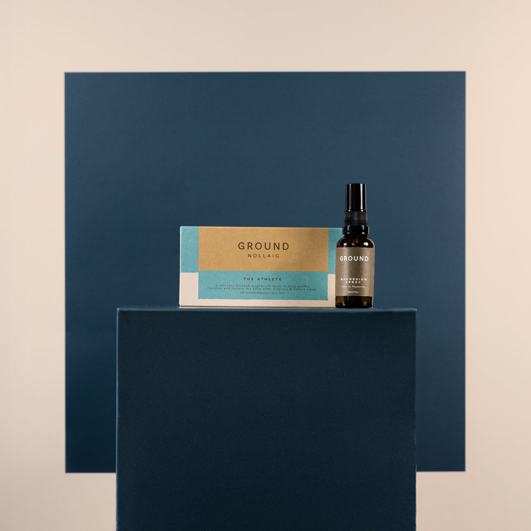 A small dark bottle of magnesium spray and its matching box labeled The athlete are displayed on a dark blue pedestal against a beige background with a matching blue geometric backdrop.