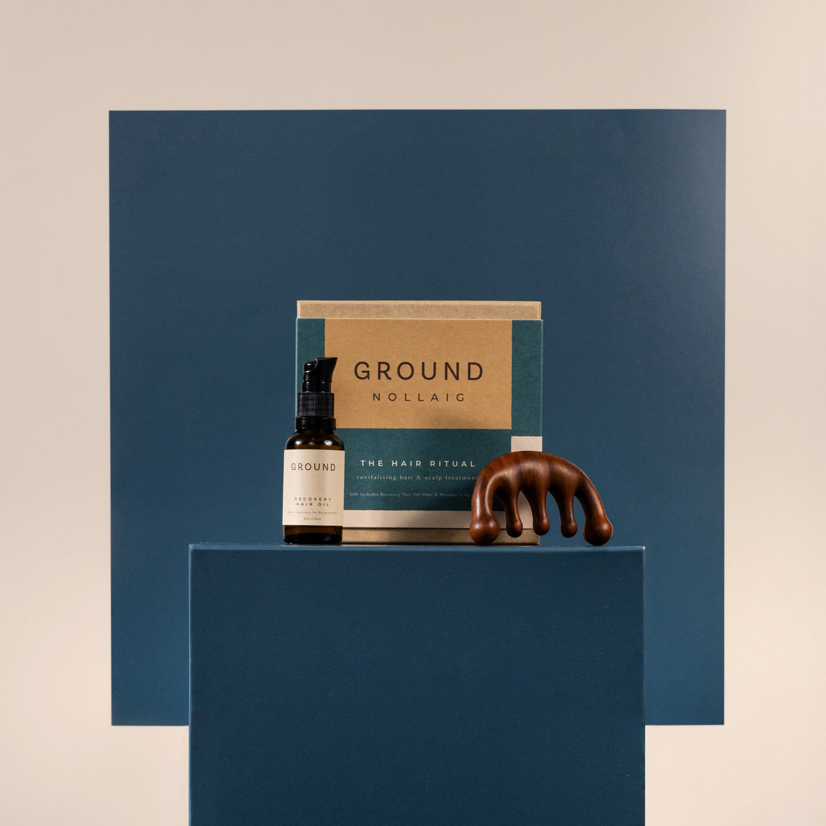 A Haircare set from Ground including a hair oil in a dark amber bottle, its kraft paper box, and a sculptural brown claw massager, displayed on a dark blue pedestal against a beige background.
