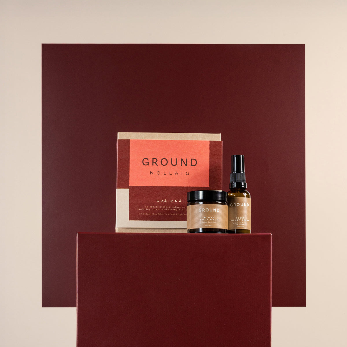 A skincare set from Ground featuring a red and beige box, a jair with night body balm and a pillow spray displayed on a dark burgundy pedestal against a light beige background.