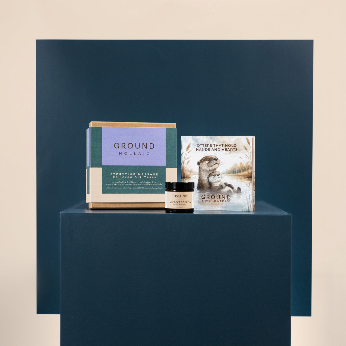 A curated wellness gift set from Ground Wellness displayed on a dark blue pedestal. The set includes a a small jar of comfort balm 60ml and a book titled 'Otters that hold hands and hearts' against a matching dark blue backdrop.