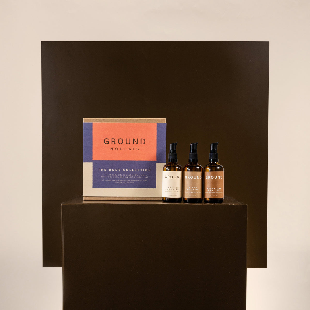 A boxed set from GROUND SCENTS called The Body Collection, displayed on a dark pedestal alongside three small amber bottles with black caps against a minimalist beige and dark brown background.