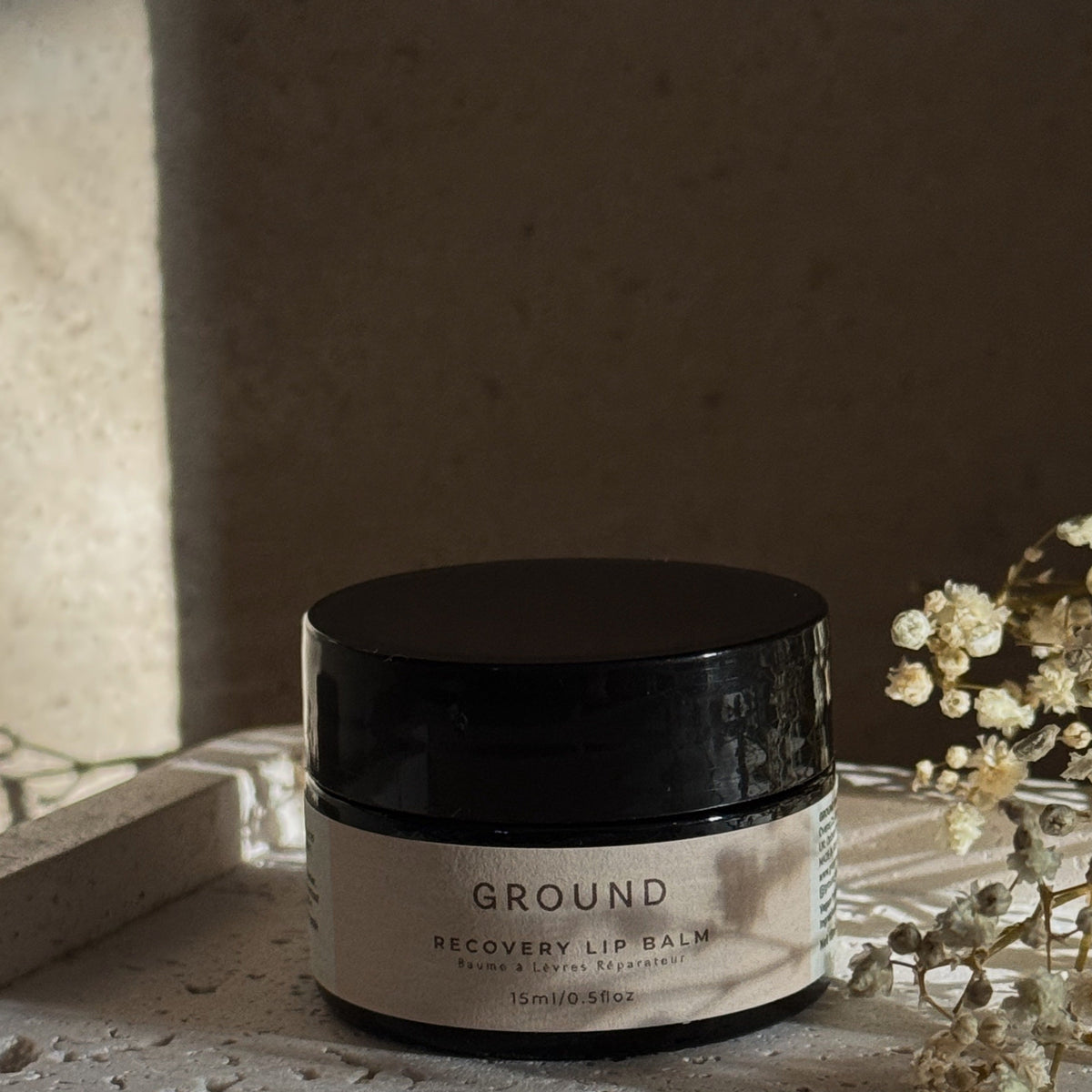 A black jar of GROUND Recovery Lip Balm sits on a textured stone surface next to dried white flowers, with warm natural light casting soft shadows.