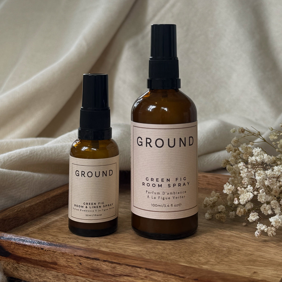 Two amber glass spray bottles with black caps and cream labels from the brand GROUND, displayed on a rustic wooden tray with neutral draped fabric and dried flowers.