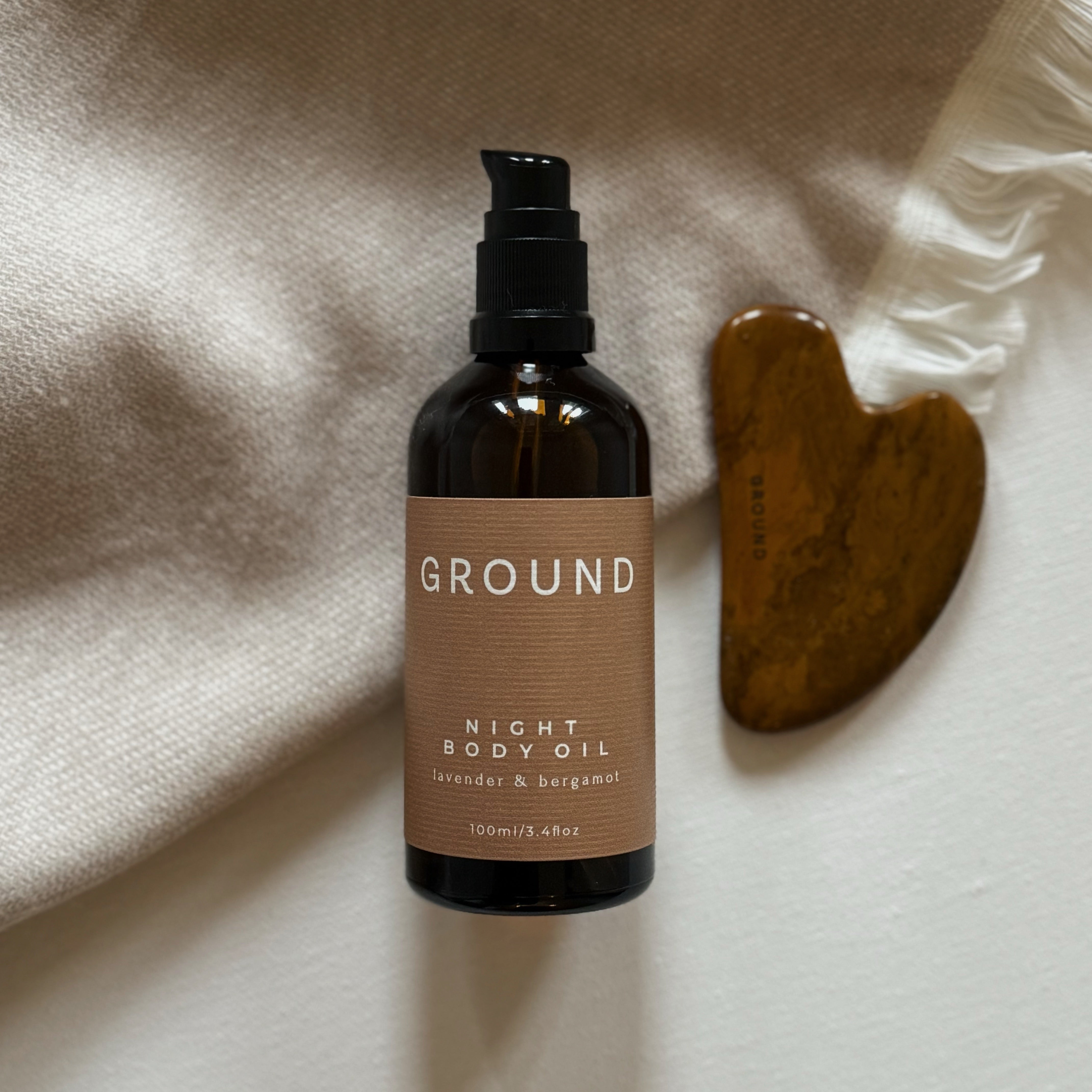 Bottle of 100ml GROUND Night Body Oil on a blanket with a gua sha