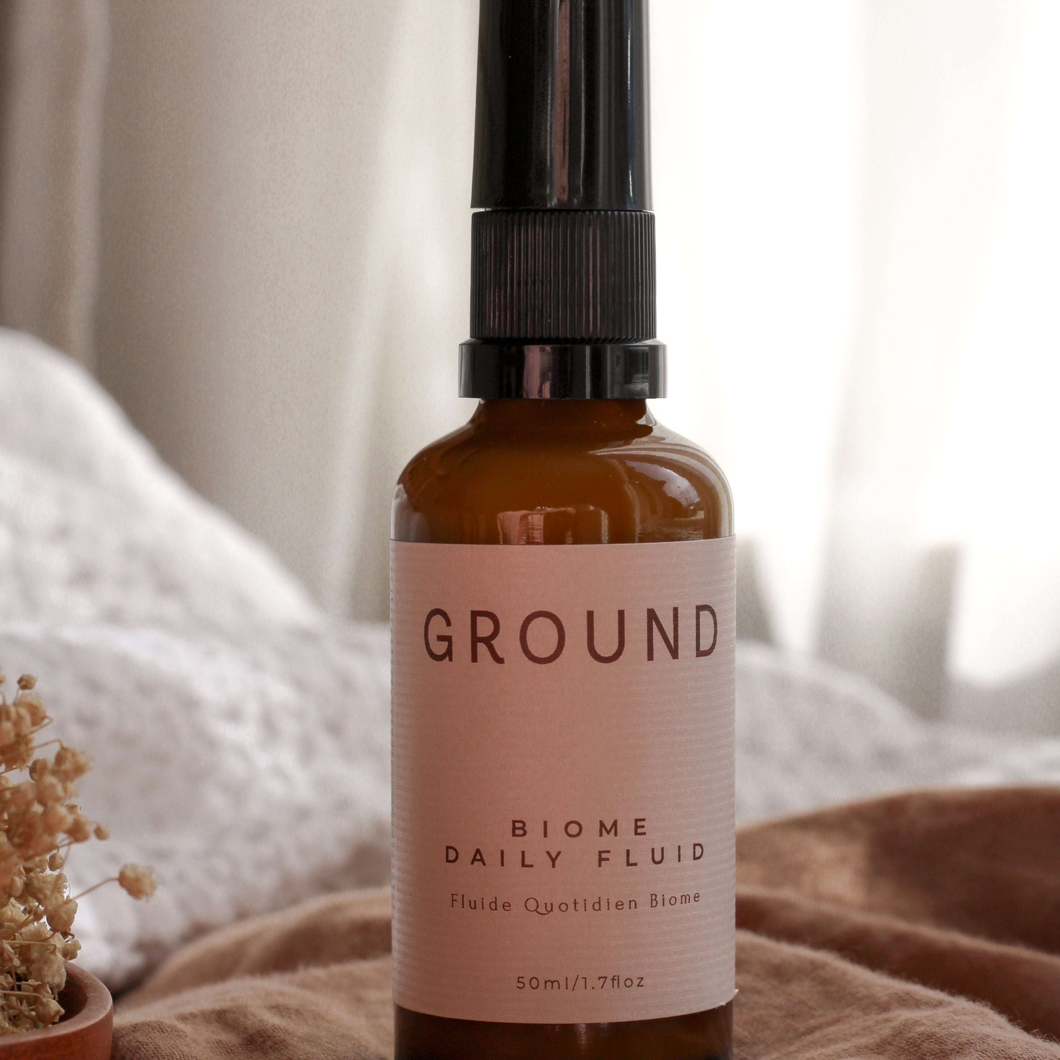 A 50ml amber glass spray bottle of GROUND Biome Daily Fluid with a black spray nozzle and a cream label, resting on a textured beige blanket with a soft, warm background.