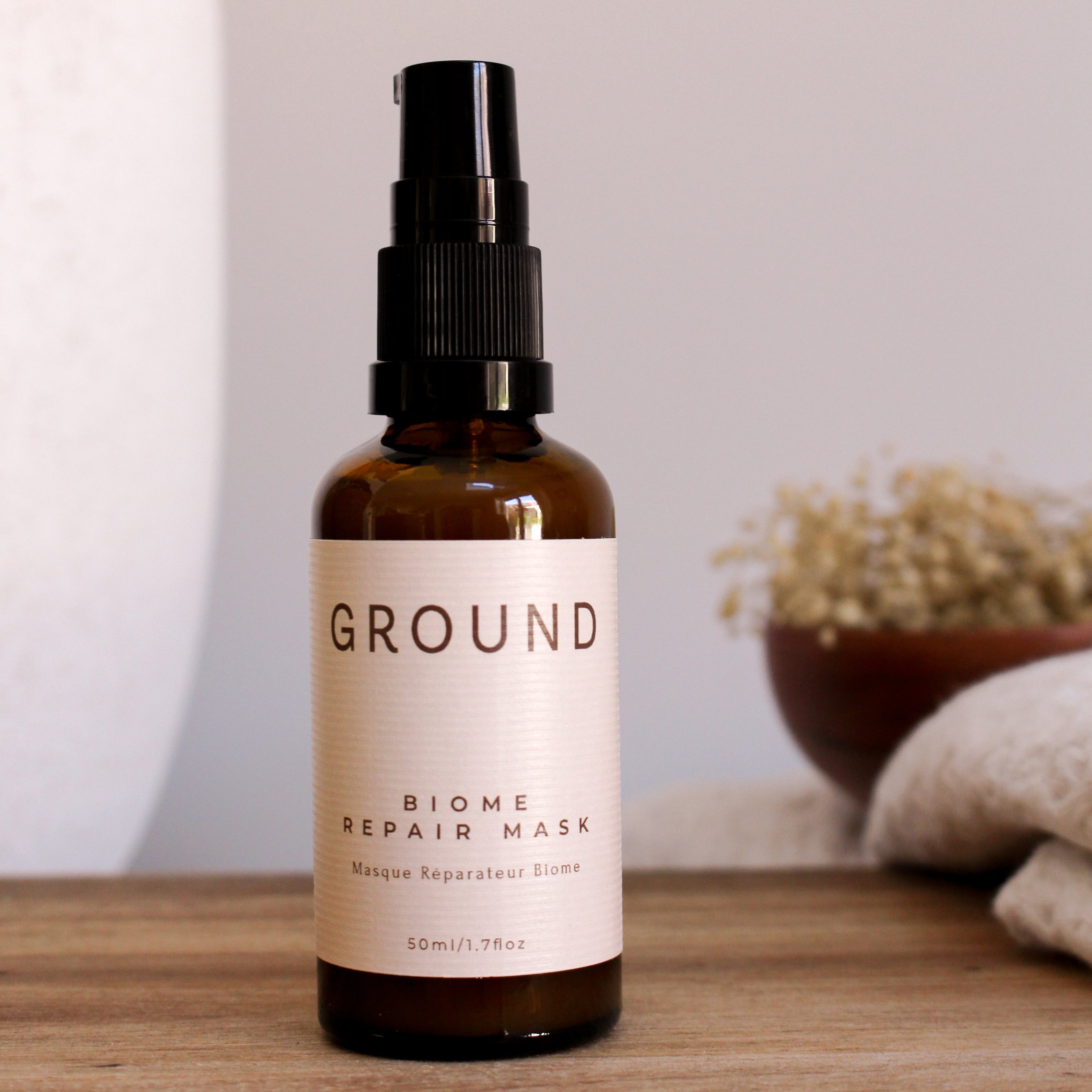 A 50ml amber glass spray bottle of GROUND Biome Repair Mask with a black pump and cream label, set on a wooden surface with a blurred neutral background.