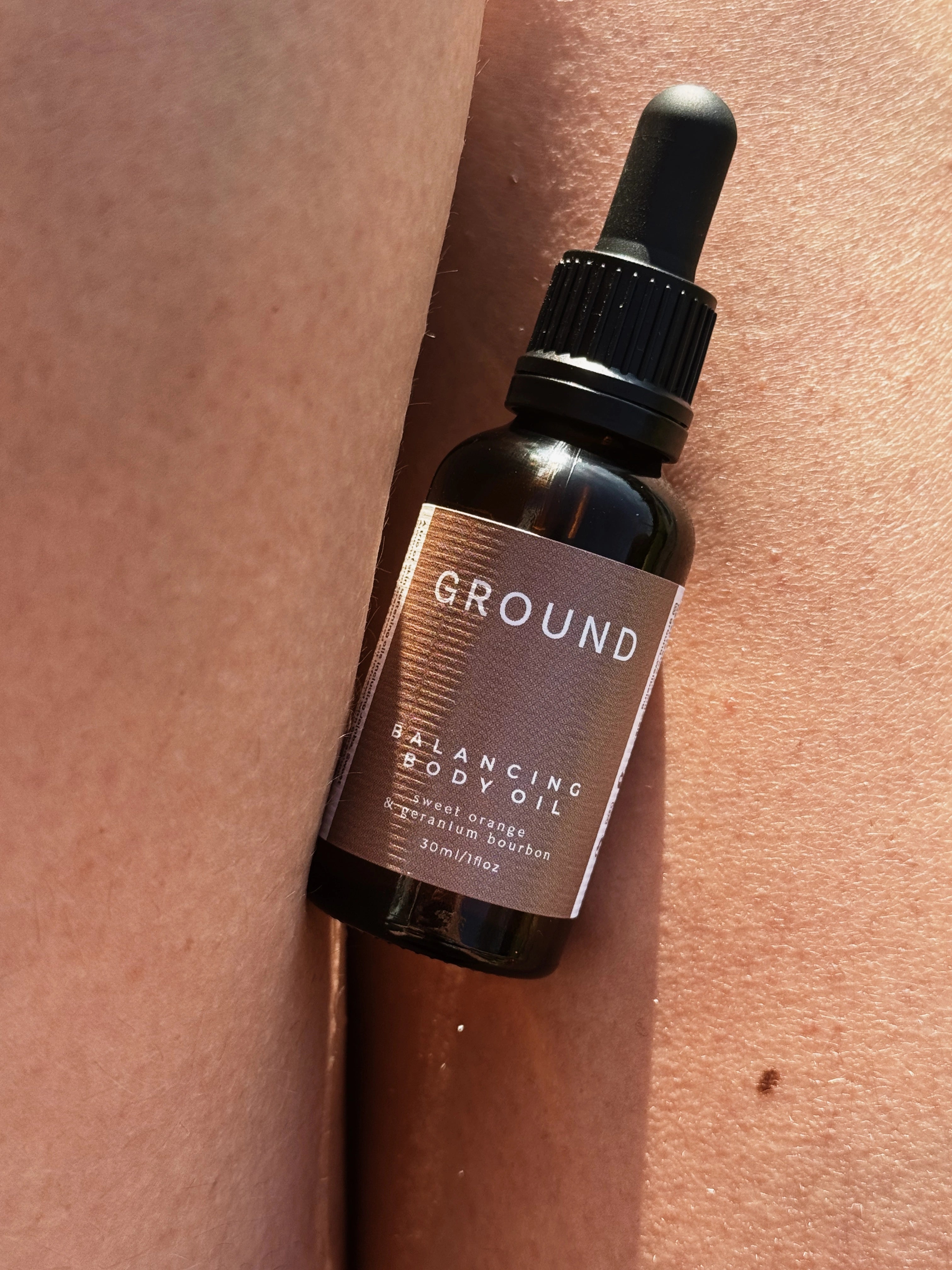 Image of brown glass bottle 30ml in size with a black dropper lid. It has a neutral brown label describing the contents as Ground Balancing Body Oil - sweet orange, geranium, bourbon - 30ml / 1 fl oz.