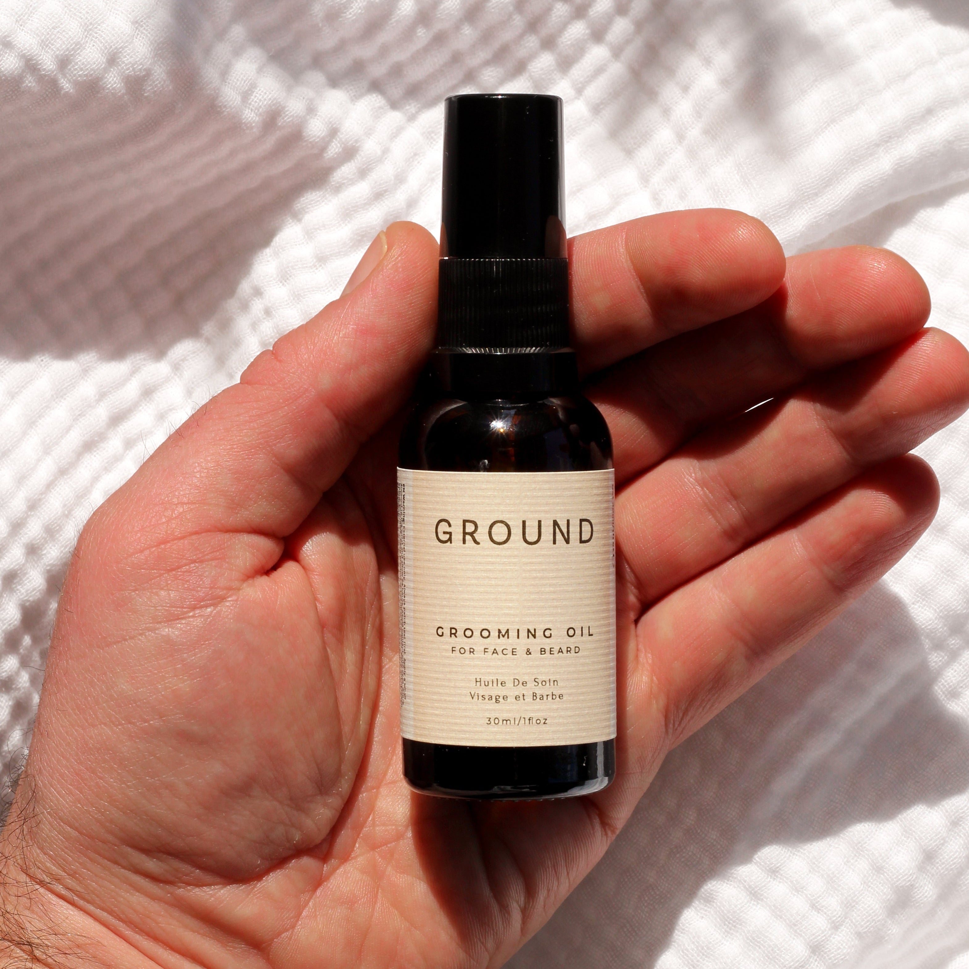 Hand holding a bottle of 'GROUND' grooming oil for face and beard against a white textured background.