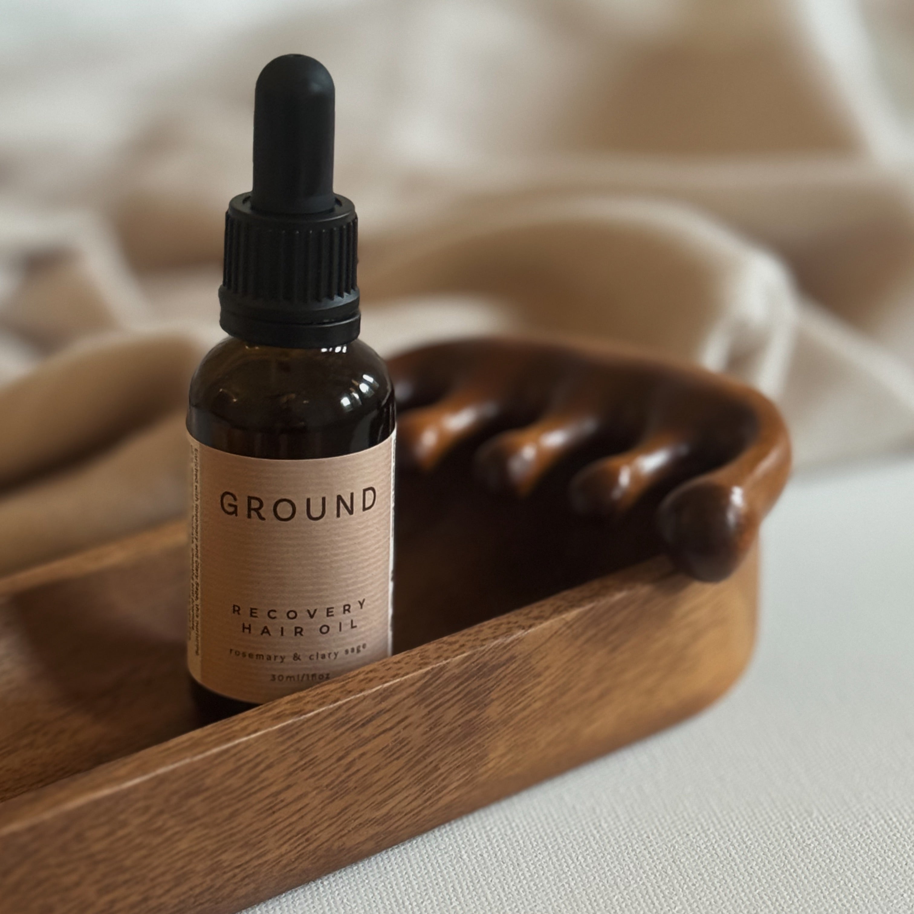 Bottle of 'GROUND' recovery hair oil on a wooden tray with a wide toothed wooden comb. The background contains beige material but is has been blurred. The label on the bottle state the hair oil contains rosemary and clary sage.