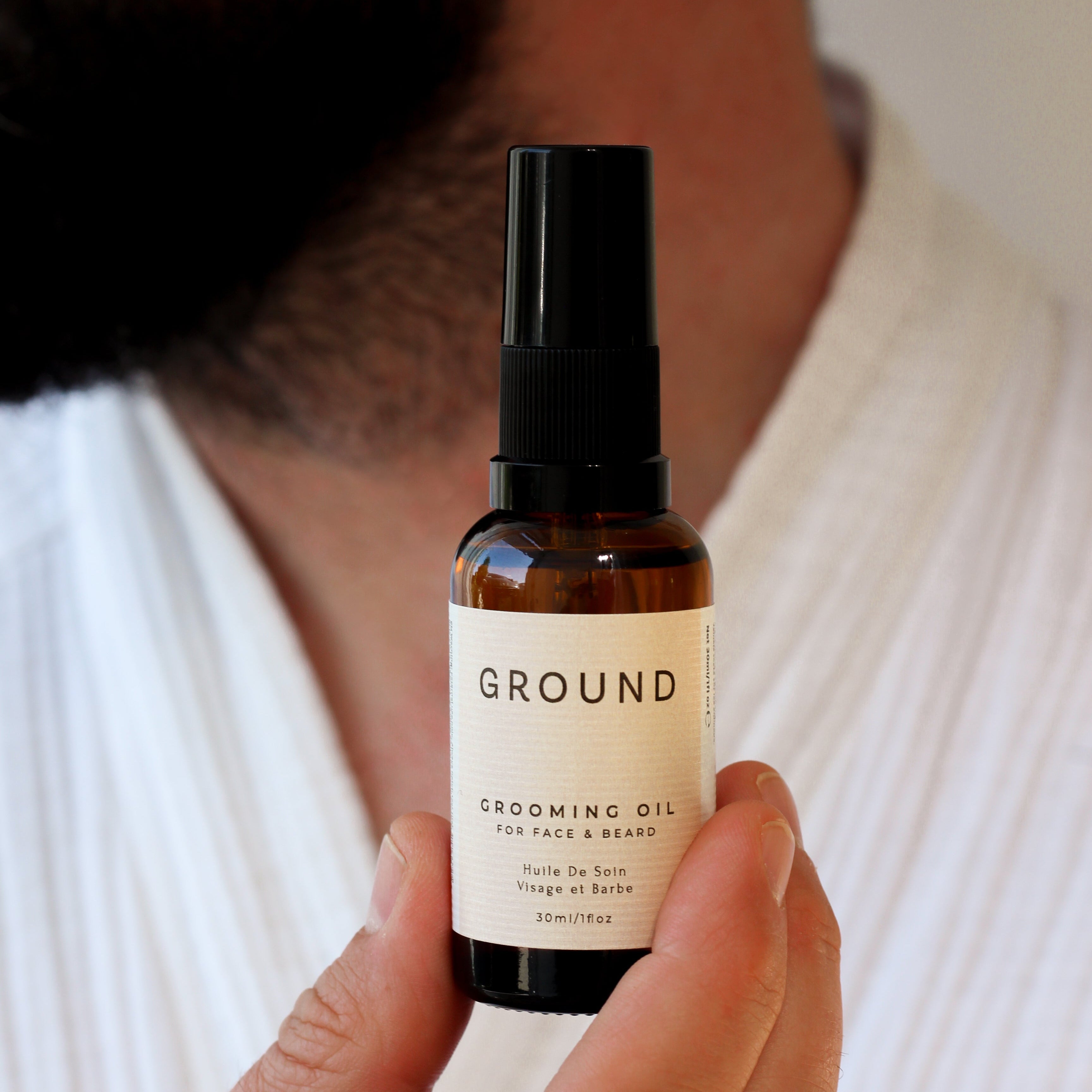 Hand holding a brown glass bottle of Ground Grooming Oil for face and beard. Held up in mid air in front of a black beard and a white top.