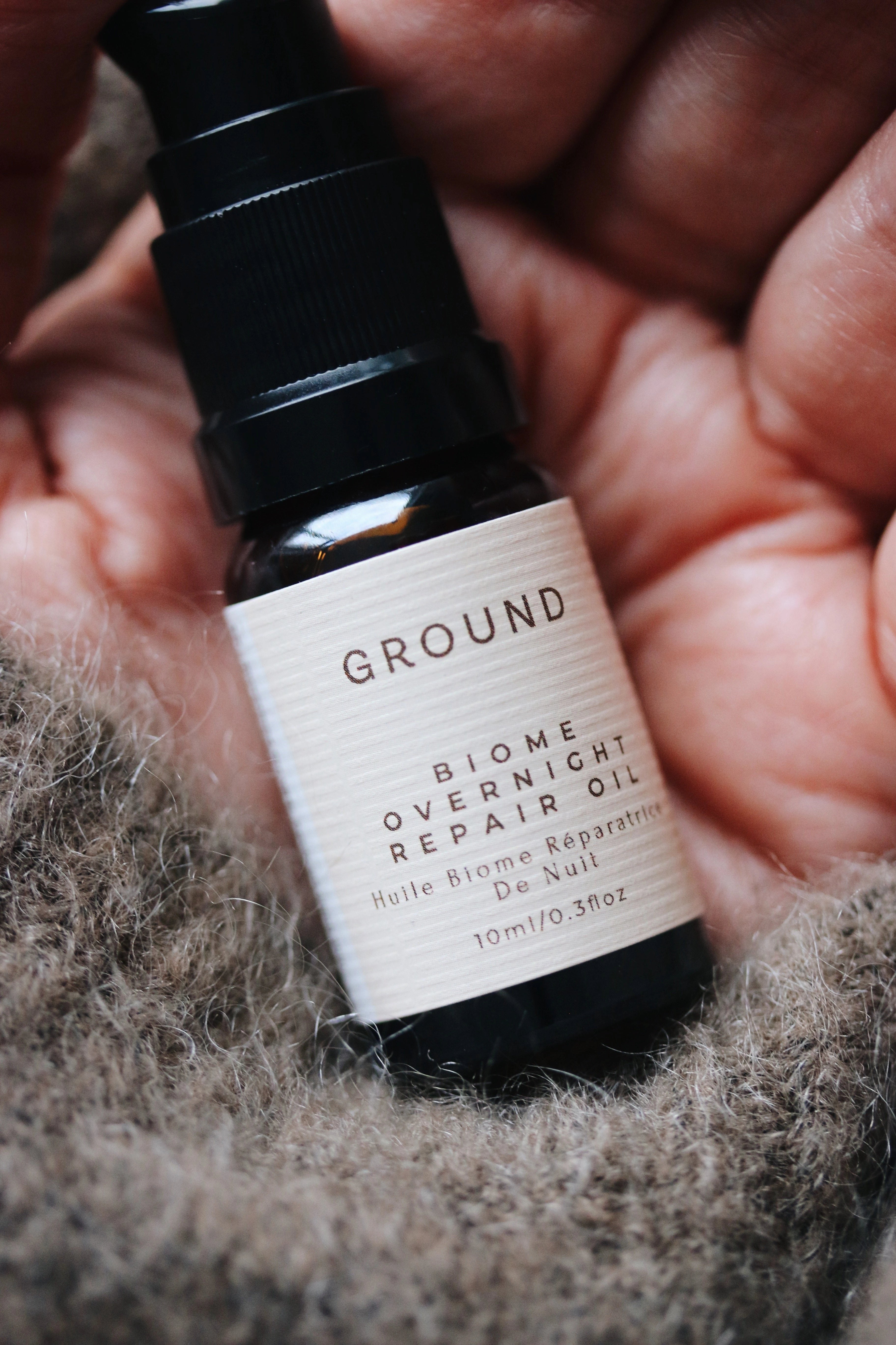 Photograph of a small bottle of Ground Biome Overnight Oil being held in a hand showing the sleeve of a grey mohair jumper or cardigan.