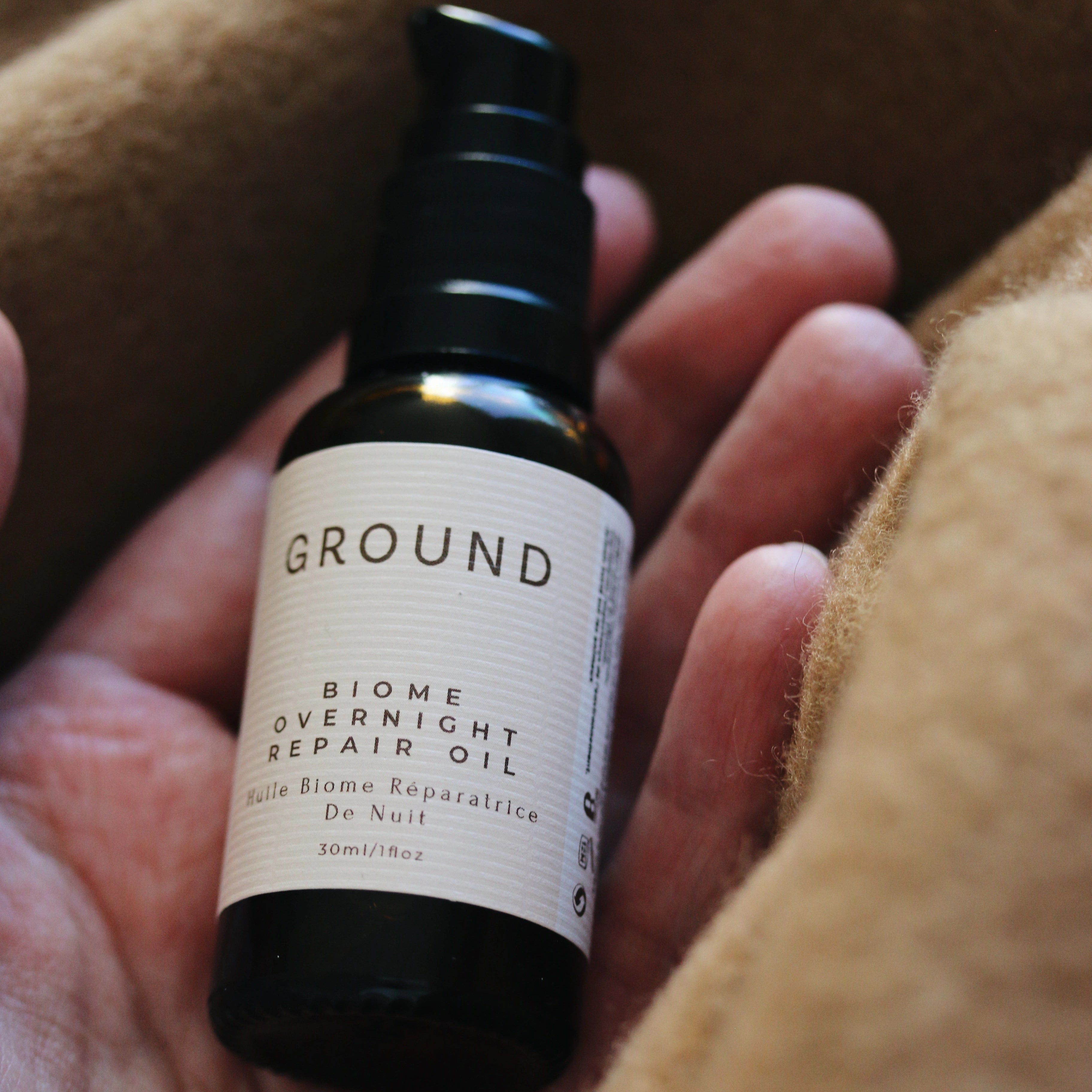 Image of a small bottle of Ground Biome Overnight Oil being held in a hand surrounded by camel beige coloured material. 