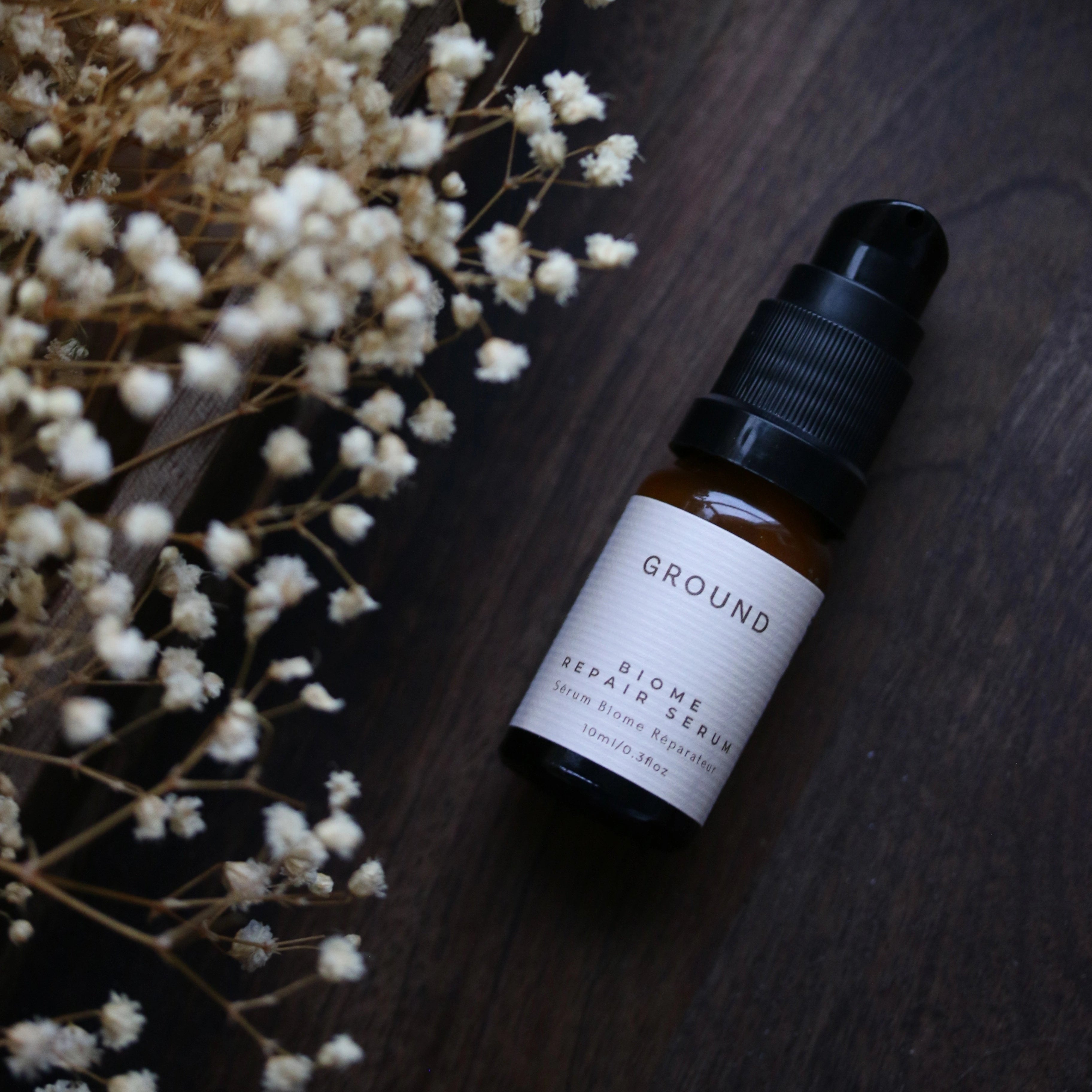 Photograph of a 10ml bottle of Ground Biome Repair Serum - Serum Biome Reparateur. It is lying flat on a dark wood surface with baby's whisper small white flowers laying next to it on the left hand  side.