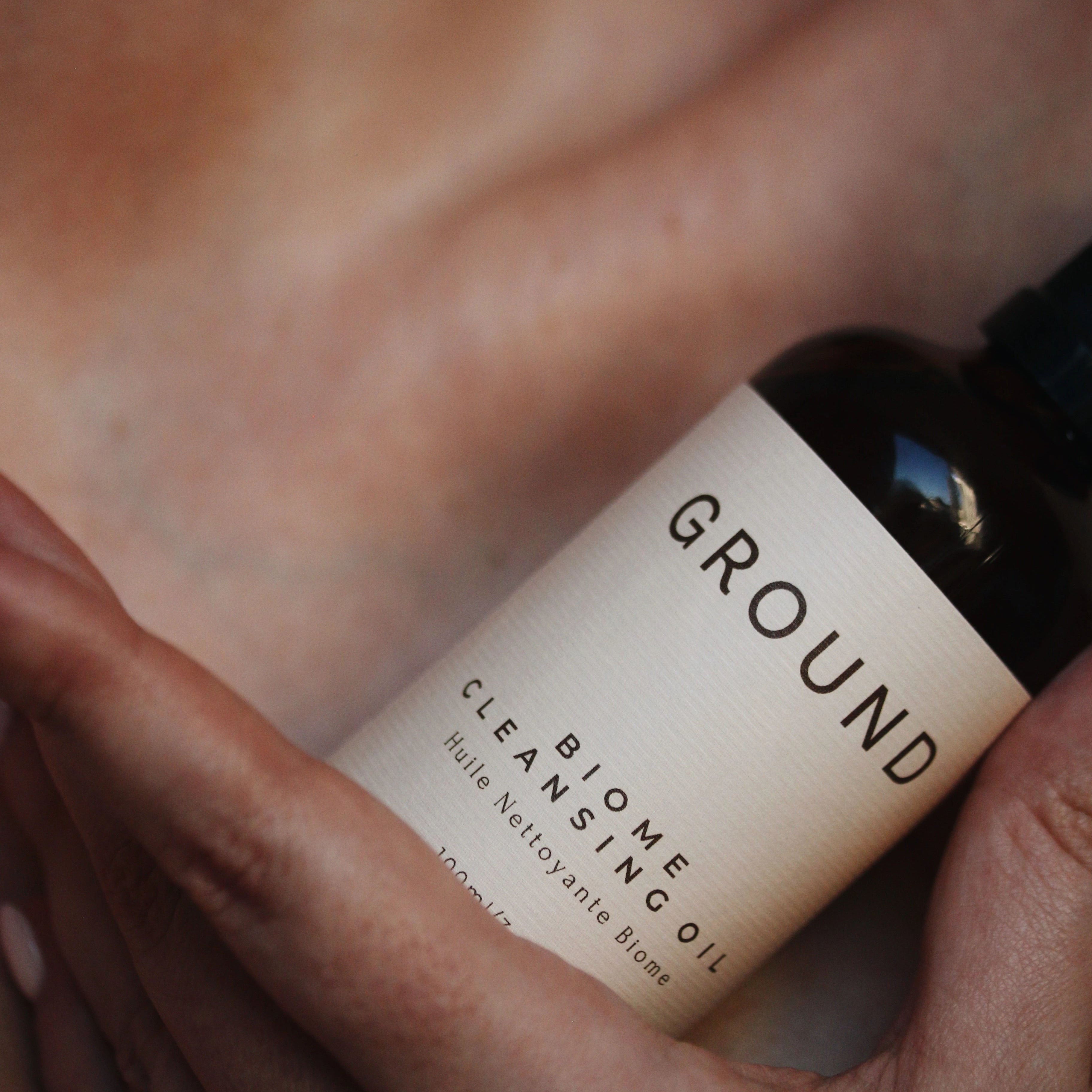 Photo of a 100ml bottle of Ground Biome Cleansing Oil being held against skin at a collarbone.