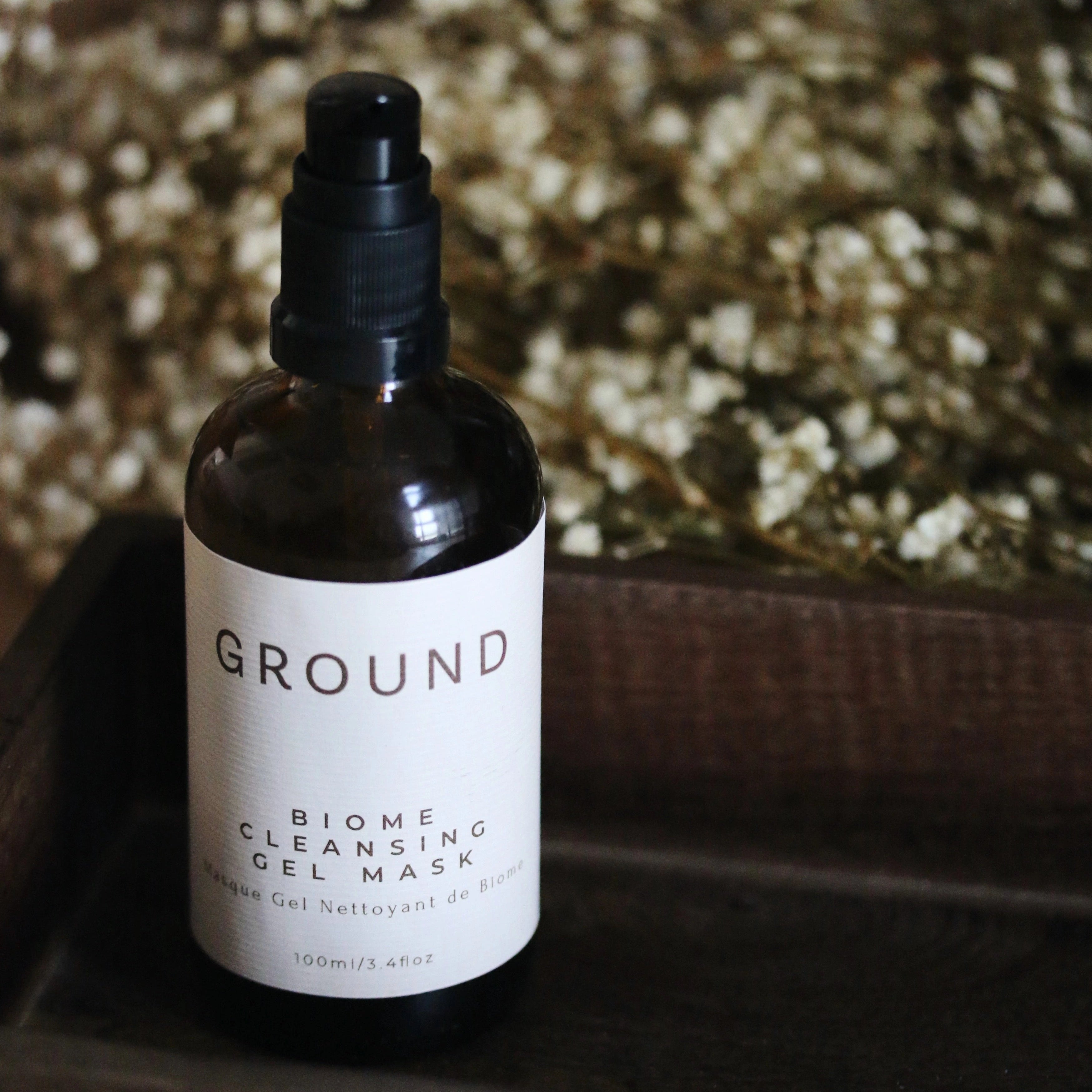 A bottle of BIOME Cleansing Gel Mask by GROUND SKIN positioned in a dark wood box, with blurred white flower buds in the background. The label is beige in colour with brown writing stating GROUND Biome Cleansing Gel Mask - 100ml.