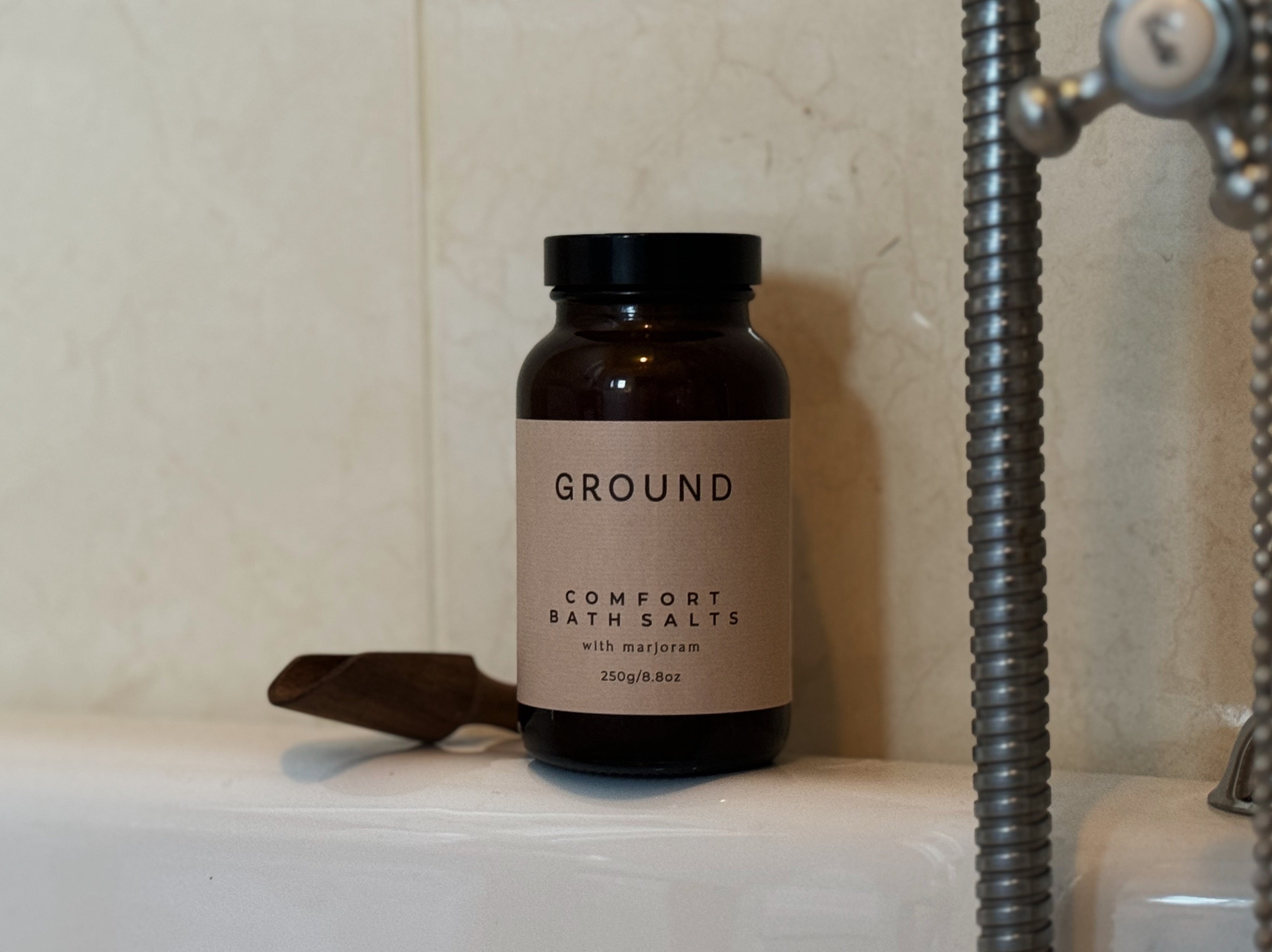 Photo of a glass bottle of Ground Comfort Bath Salts with marjoram sitting on the edge of a bath with a small scoop beside it. 
