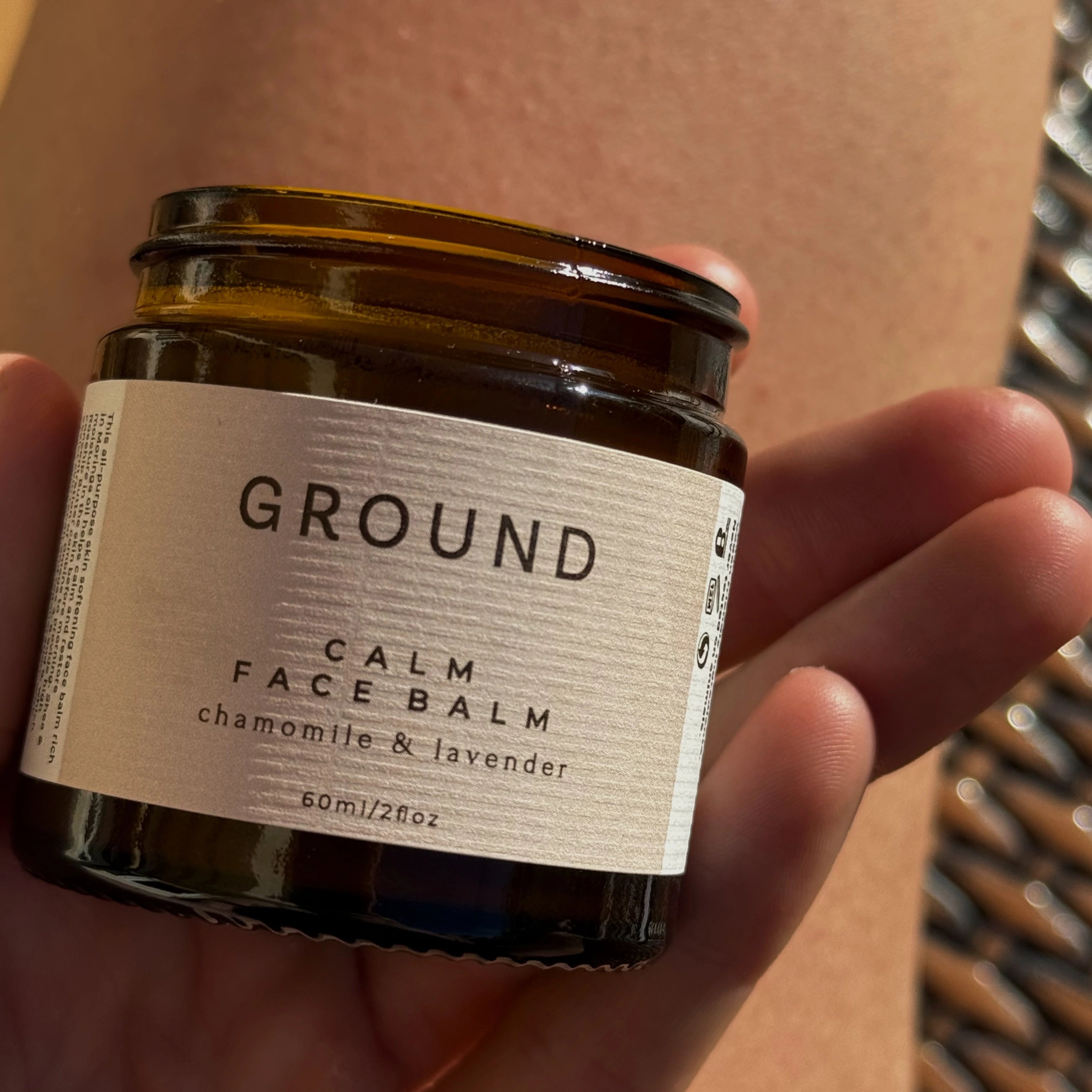 A jar of Calm Face Balm by Ground Wellbeing being held in the fingertips in a bright location. This balm contains chamomile and lavender.