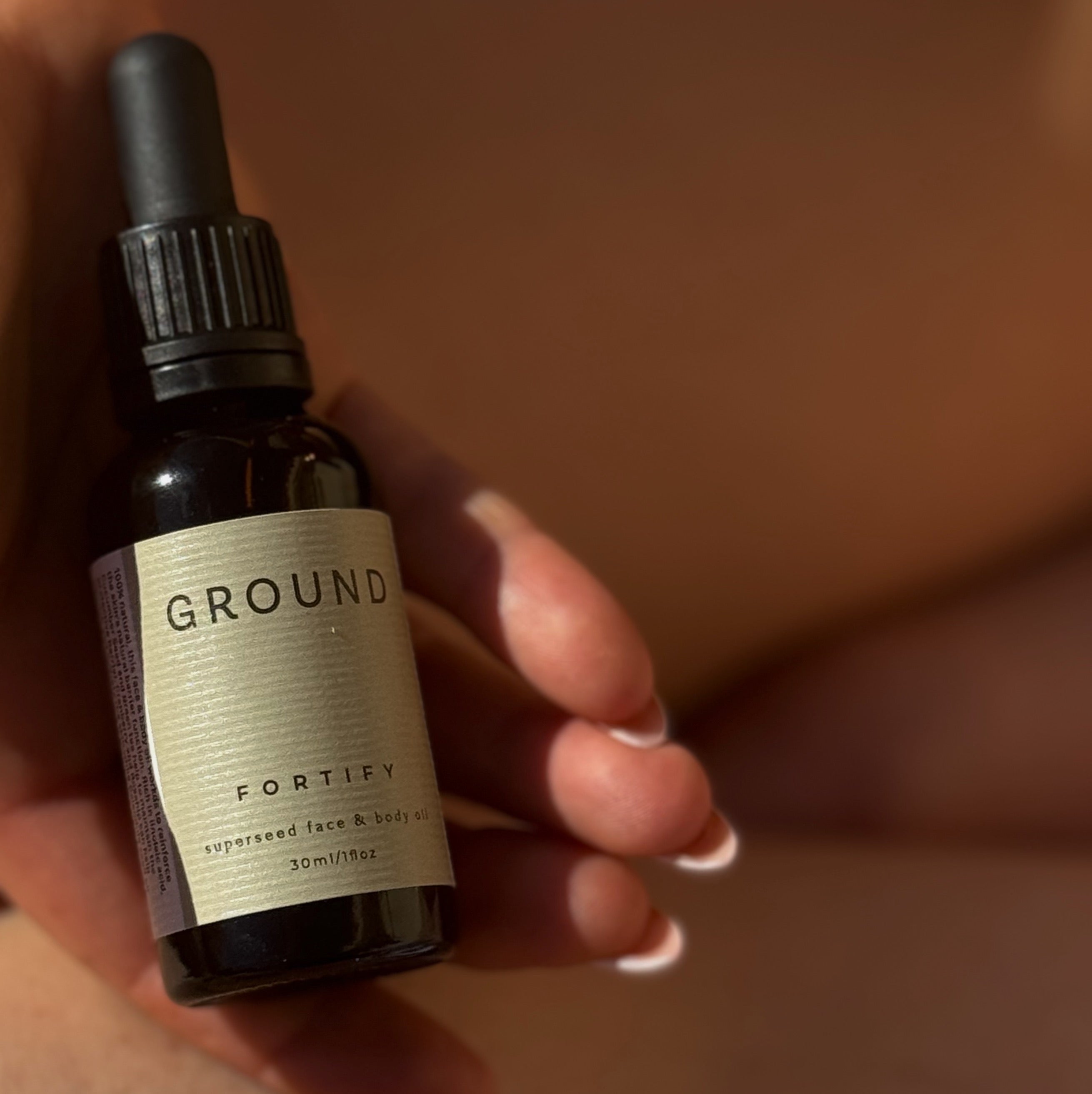 Photograph of a glass dropper bottle being held up in the sunshine. It is labelled with a sage green label stating Fortify Superseed Face & Body Oil 30ml.