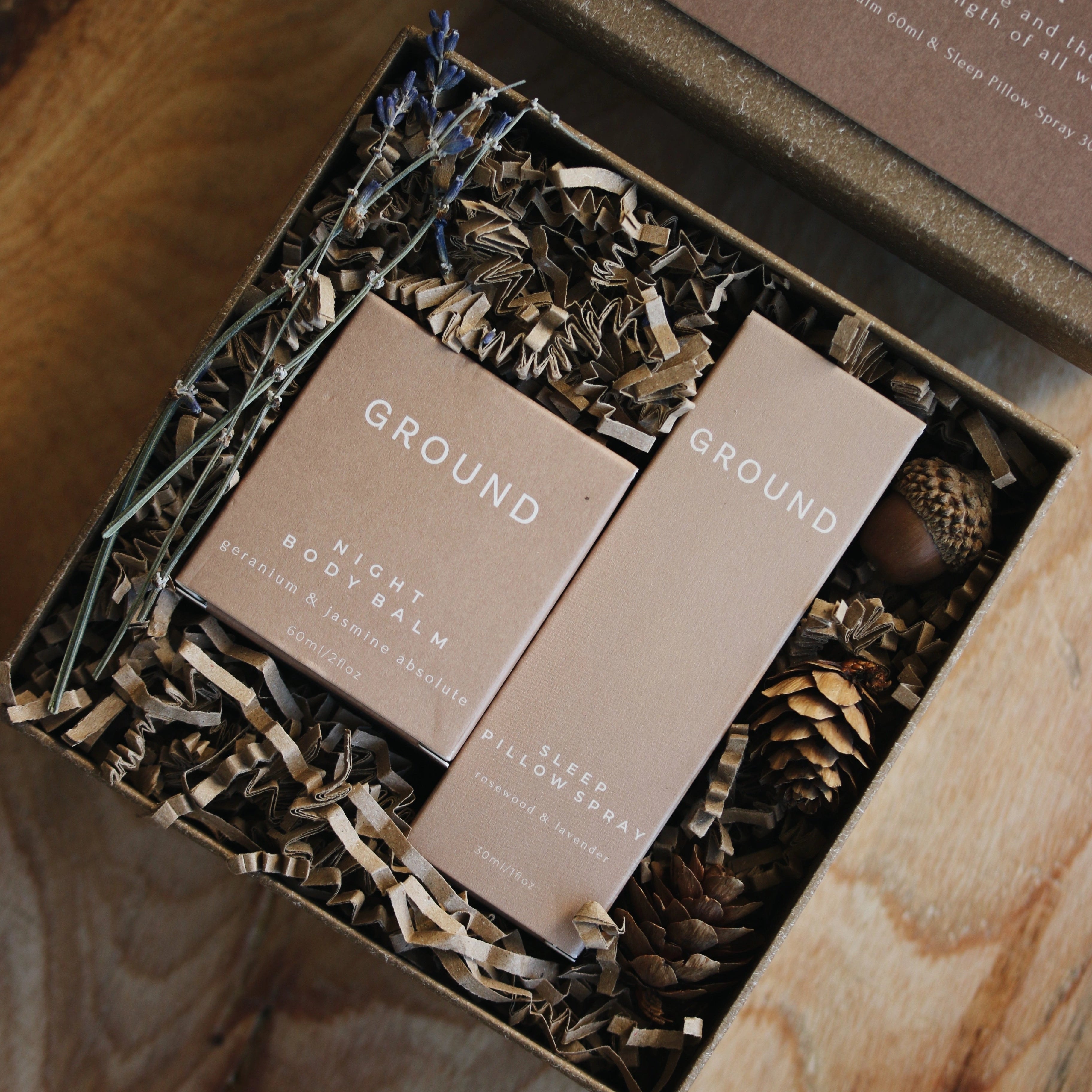 Photograph of Ground Gra Mna gift set with the lid off. It contains a Night Body Balm 60ml in a cube cardboard box, and a Sleep Pillow Spray in a rectangular cardboard package. These are inset into shredded cardboard with some sprigs of lavender and an acorn.