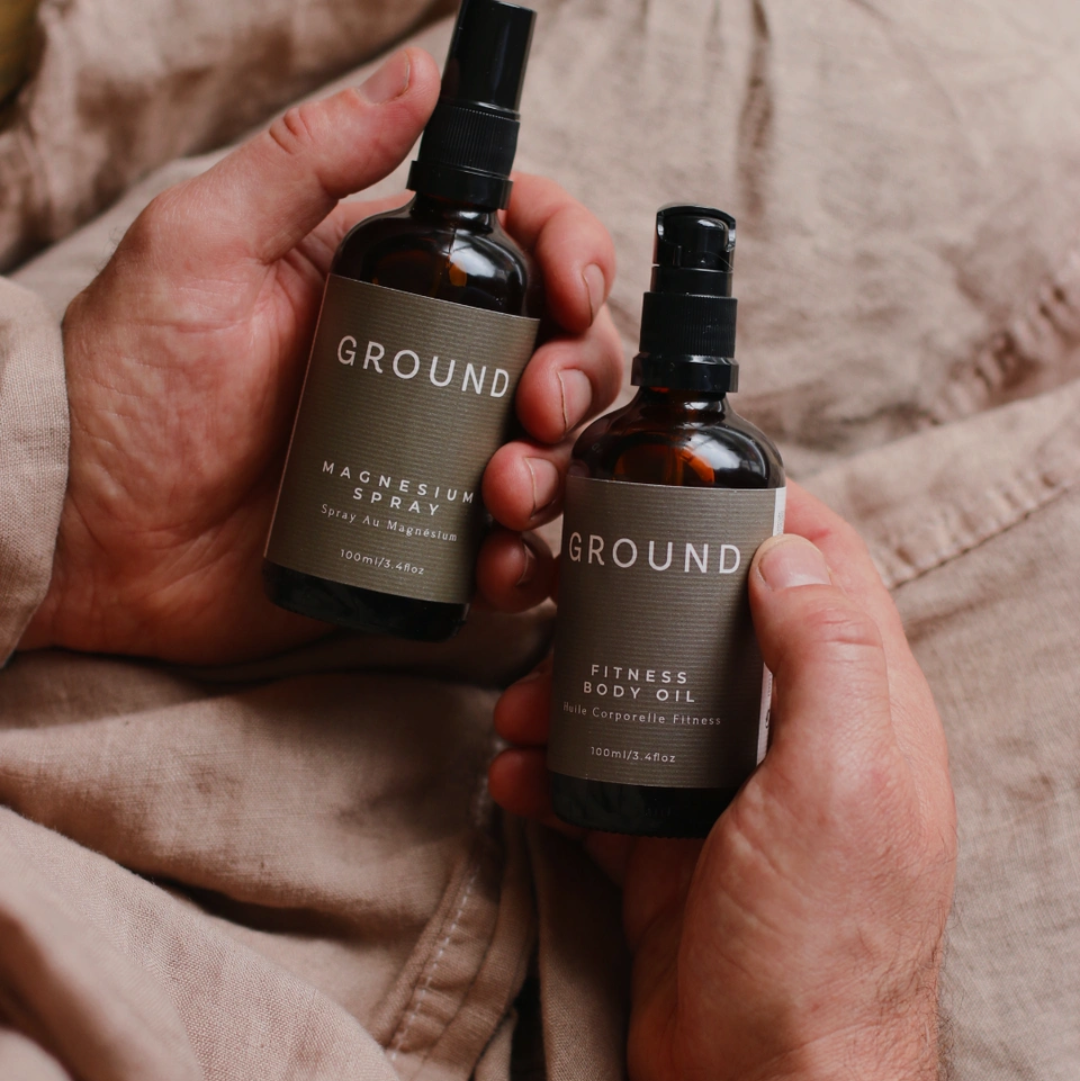 Two bottles of 'GROUND' products held by a person against a neutral linen background. One is Ground Magnesium Spray 100ml and the other a Fitness Body Oil 100ml / 3.4 fl oz.