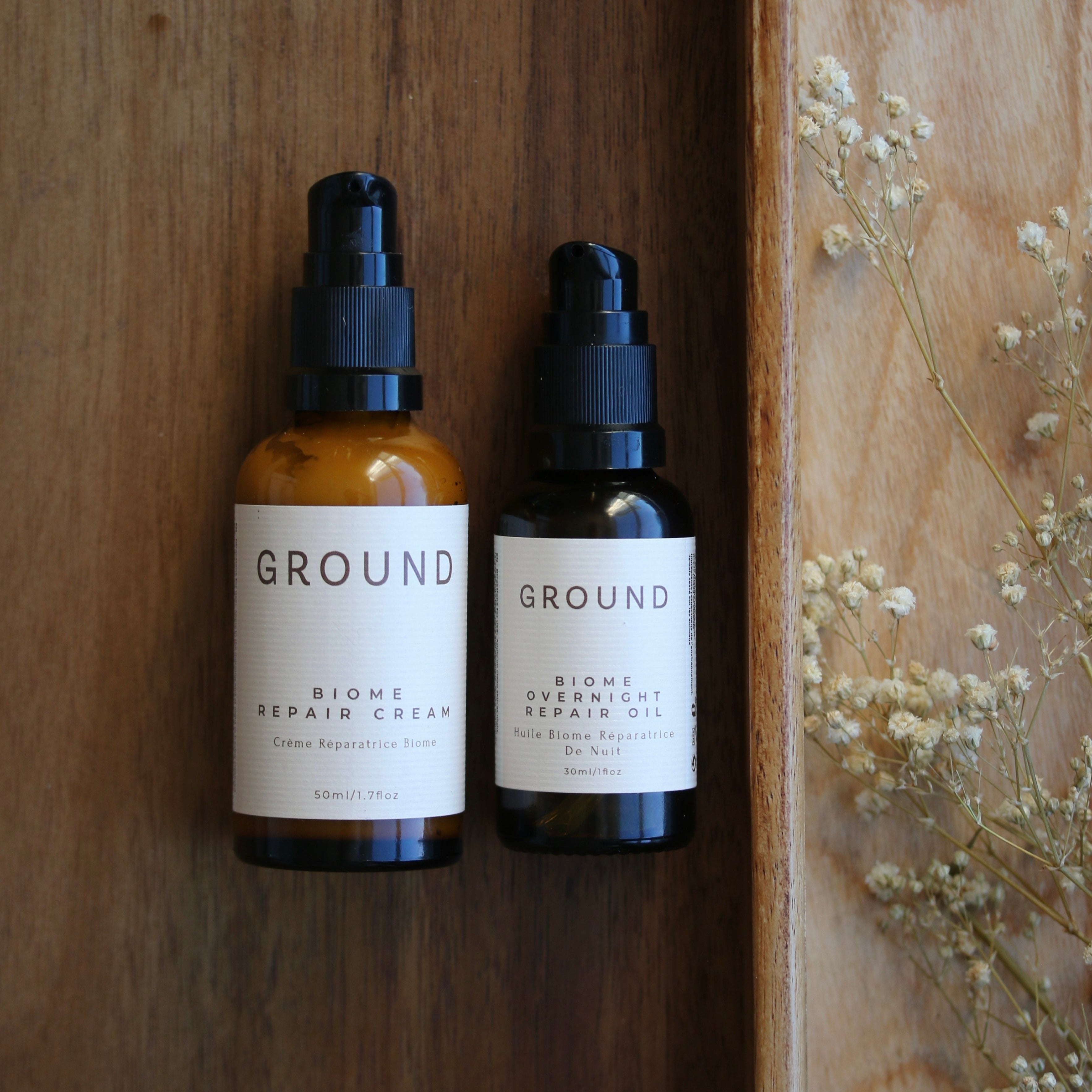 Photo shows two 'GROUND' skincare products on a wooden surface with dried flowers. One is Ground Biome Repair Cream (50mls) and the other Biome Overnight Repair Oil (30mls). Each has a pump top.
