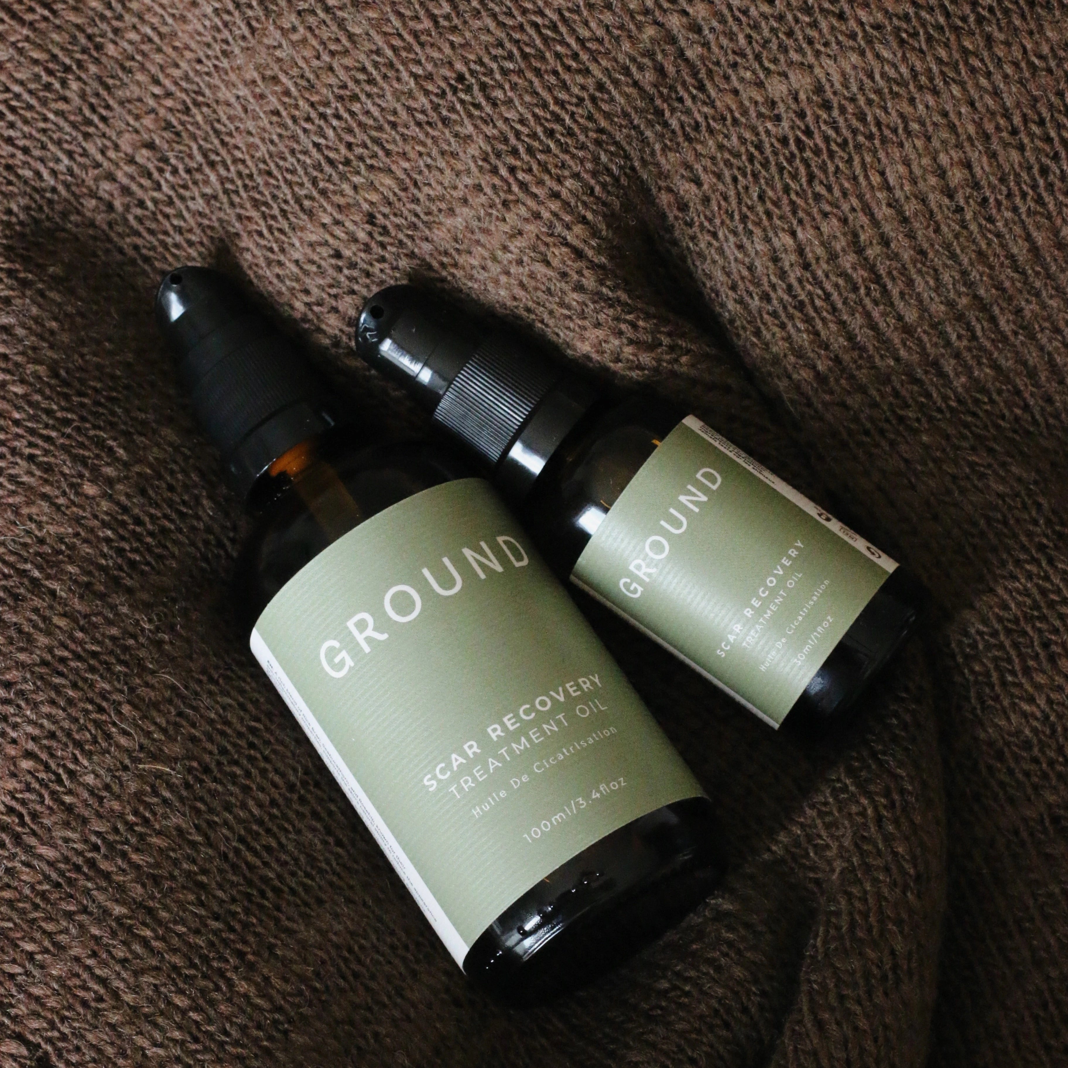 Two bottles of 'GROUND' skin recovery oil on a brown textured surface. It is available in two sizes- 100ml and 30ml and these are both shown in the photograph. Each bottle is brown glass with a sage green label with white writing. 