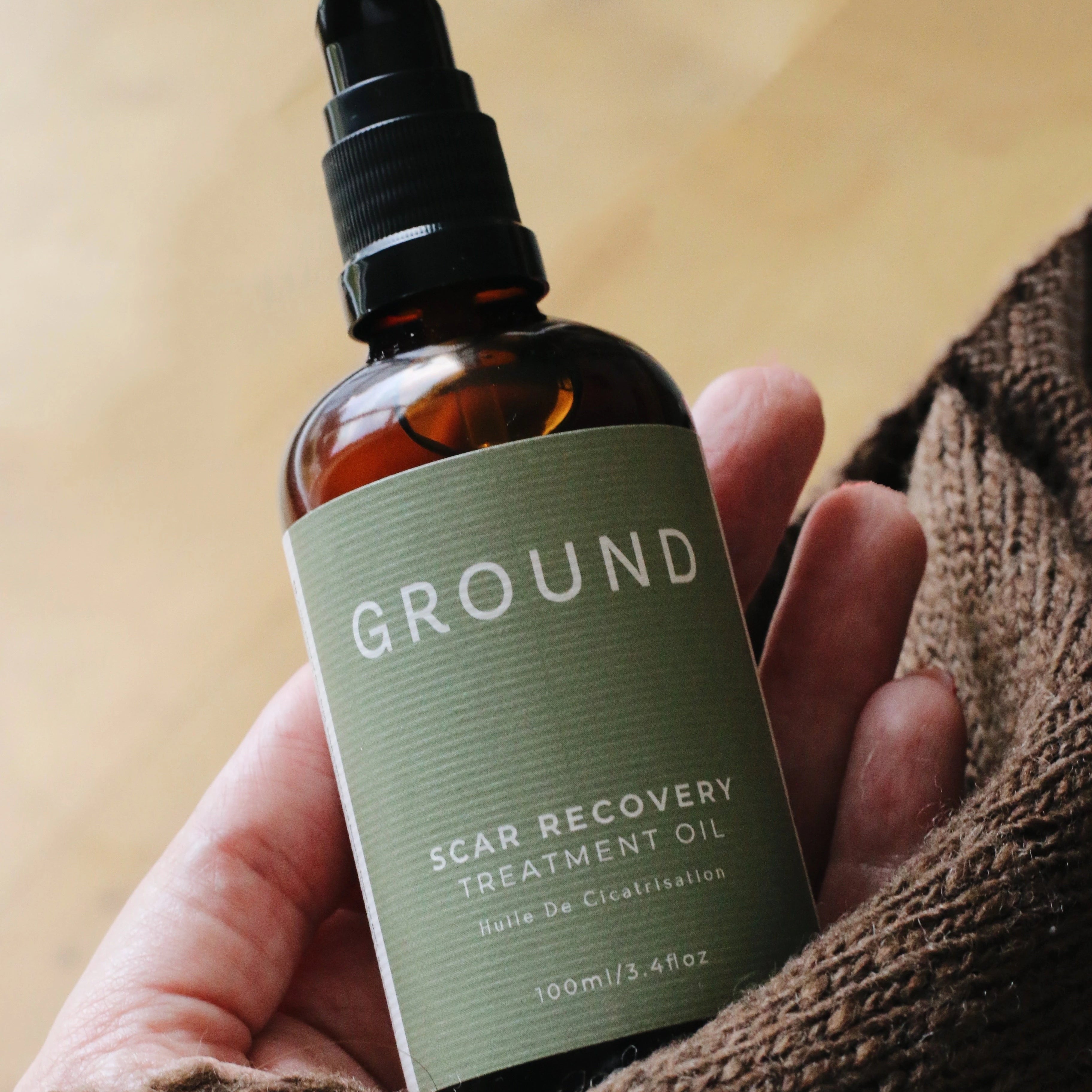 Hand holding a bottle of 'GROUND' scar recovery treatment oil with a blurred yellow background and brown textured material. This image reinforces the natural ingredients and nourishing essence of the brand - Ground Wellbeing.