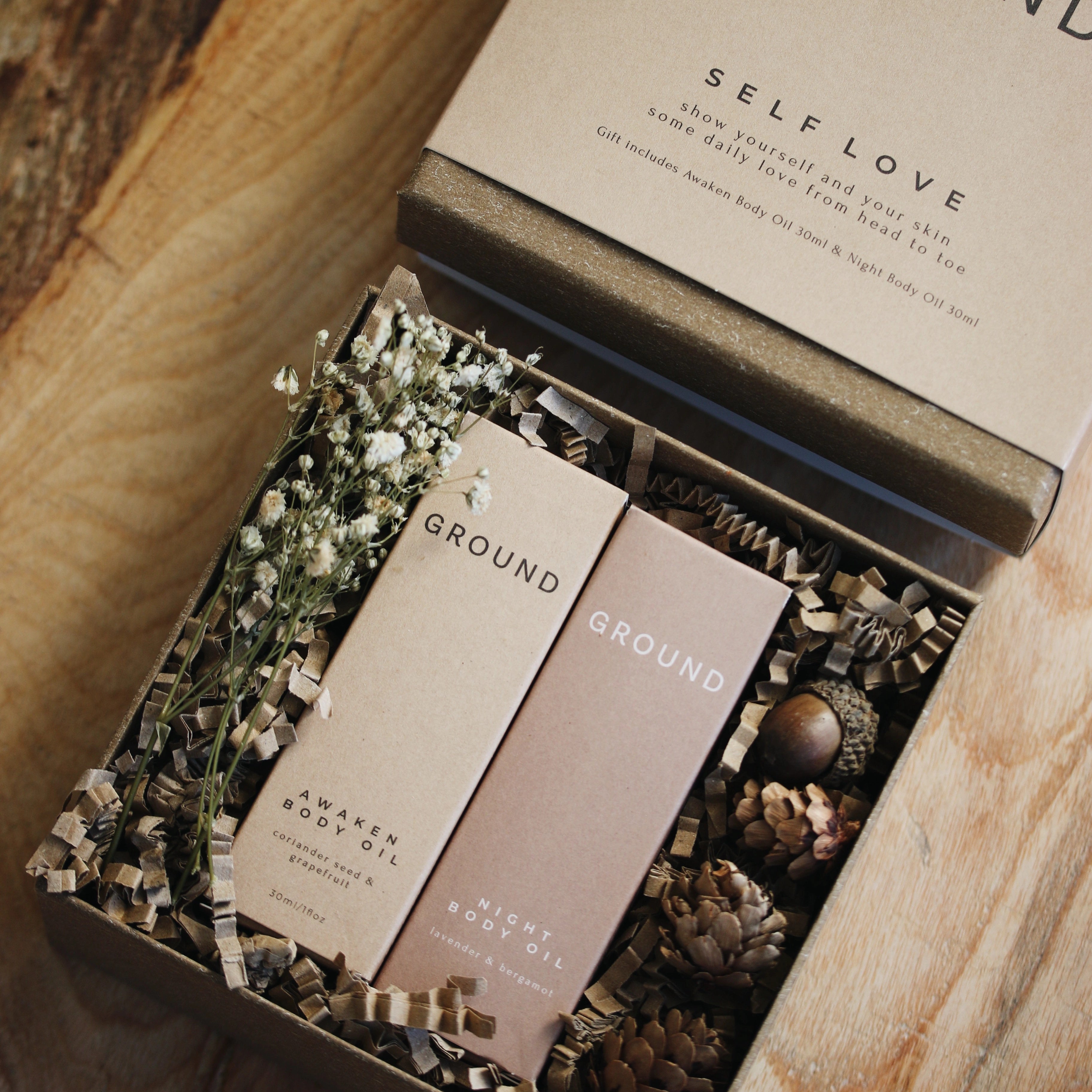 Open gift box containing two Ground Wellbeing Body Oils in their cardboard packaging, surrounded by shredded cardboard. dried white flowers, pine cones and acorns.