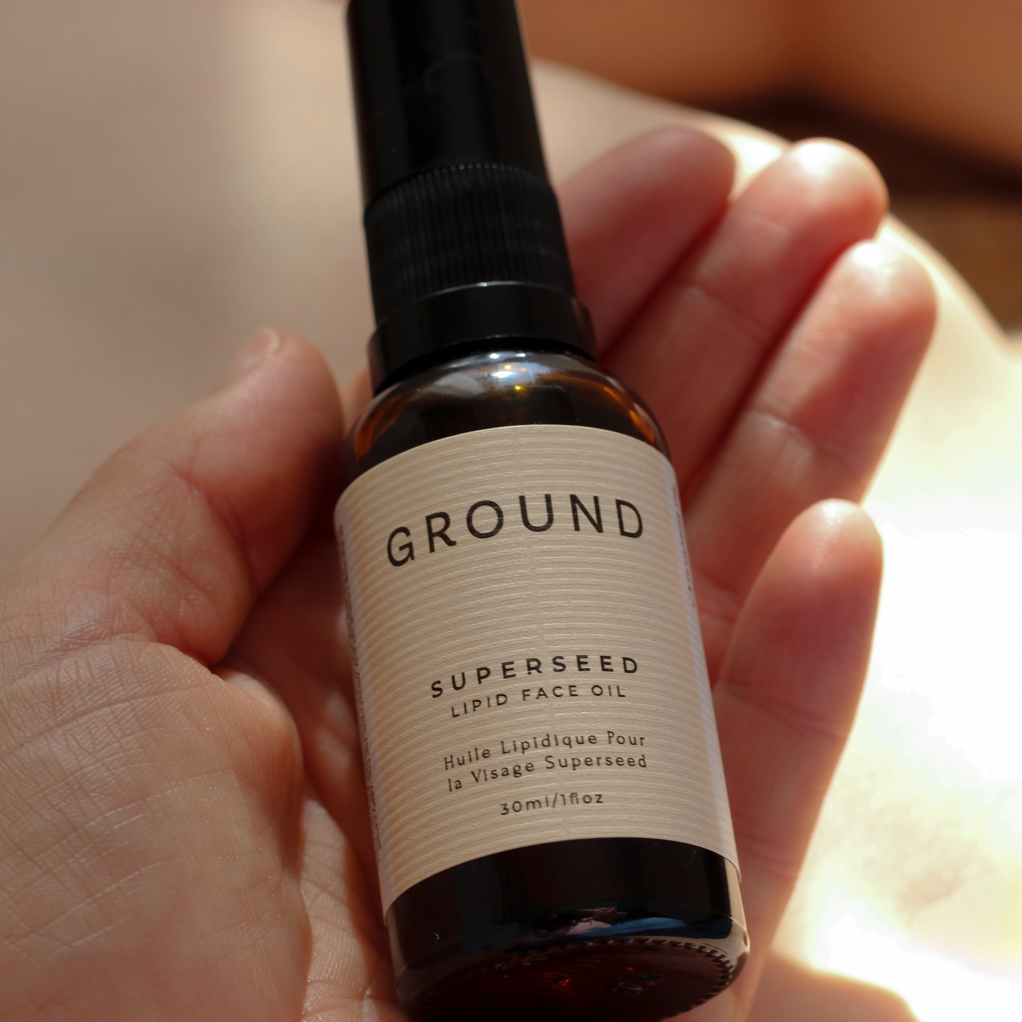 Hand holding a bottle of 'GROUND' superseed lipid face oil - Huile Lipidique Por la Visage Superseed - 30ml/1 floz. There is a warm rich peach sunlit backdrop behind the hand.
